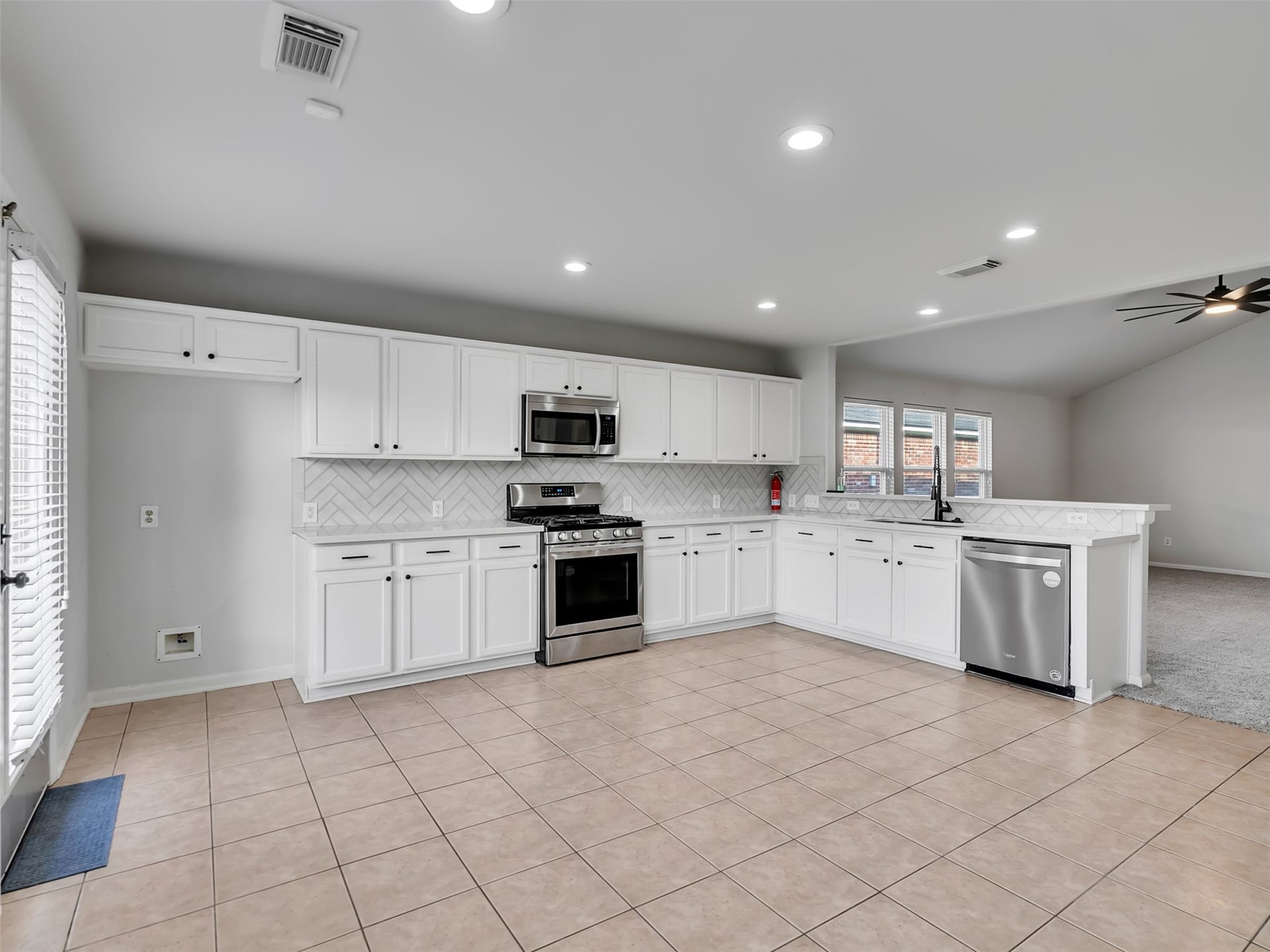 21518 Gannet Peak Way, Katy, TX 77449 This spacious kitchen features modern stainless steel appliances, white cabinetry, a stylish herringbone tile backsplash, and tile flooring. It opens to a carpeted living area with ample natural light from multiple windows.