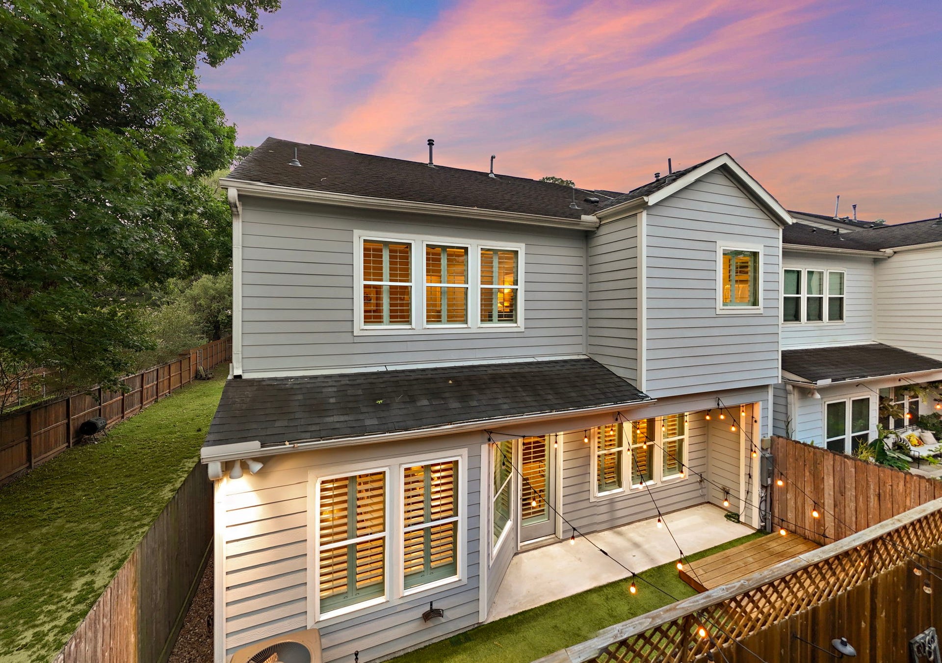 4926 Ella Boulevard, Houston, TX 77018 Charming two-story home with gray siding and white trim. The backyard features a cozy fenced area with string lights, a small lawn, and a patio, perfect for outdoor gatherings. The sunset sky adds a warm ambiance.
