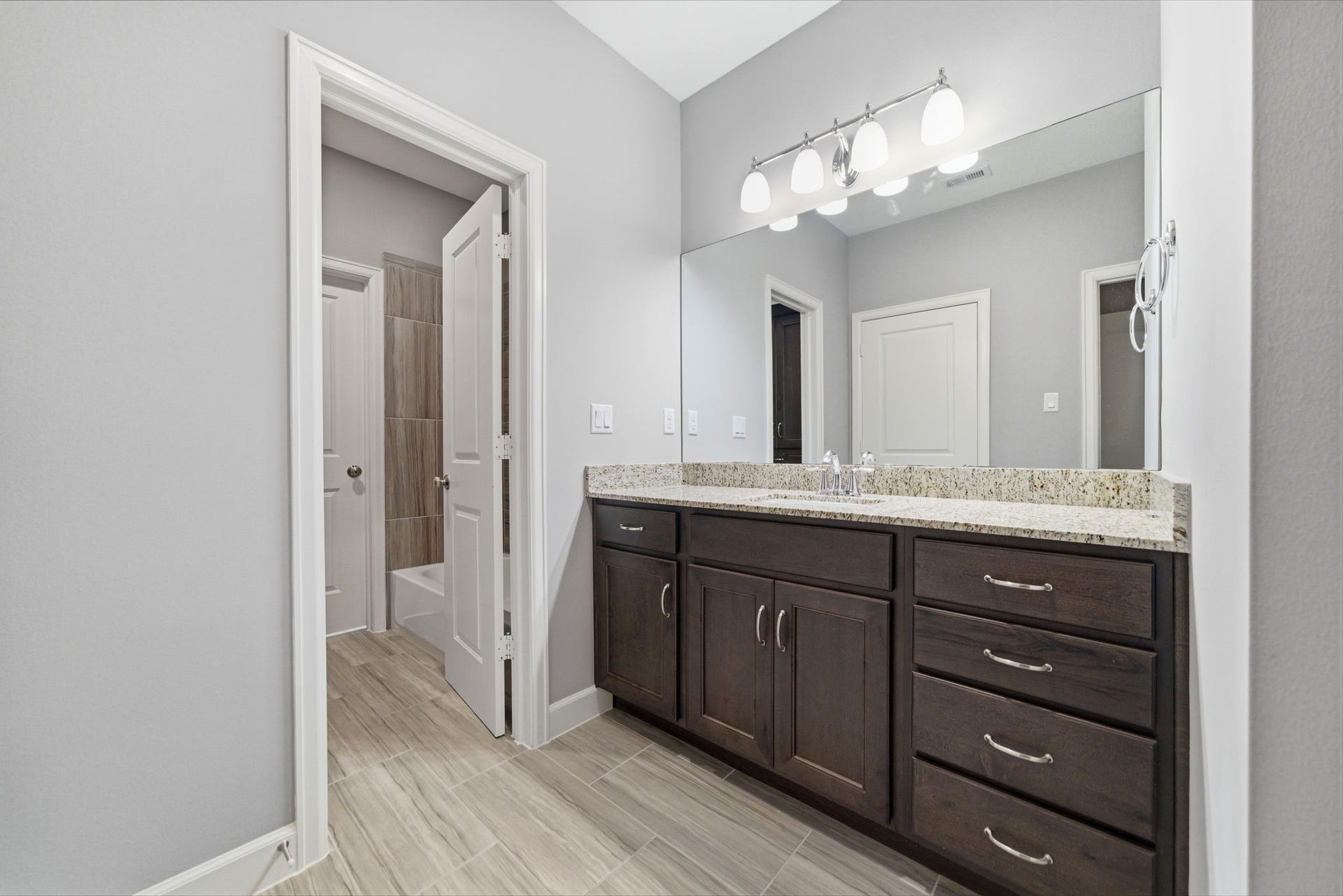 4926 Ella Boulevard, Houston, TX 77018 Guest bathroom features a granite countertop with dual sinks and dark wood cabinetry, complemented by a large mirror and modern light fixtures. The neutral tile flooring leads to a connected shower area.