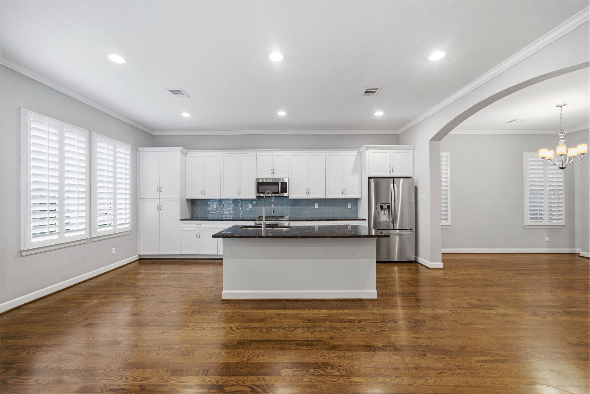 4926 Ella Boulevard, Houston, TX 77018 Spacious, open-concept island kitchen and dining area with hardwood floors, white cabinetry, a large granite island, and stainless steel appliances that includes the refrigerator. The room is brightened by recessed lighting and large windows with shutters. An elegant archway leads to the dining space.