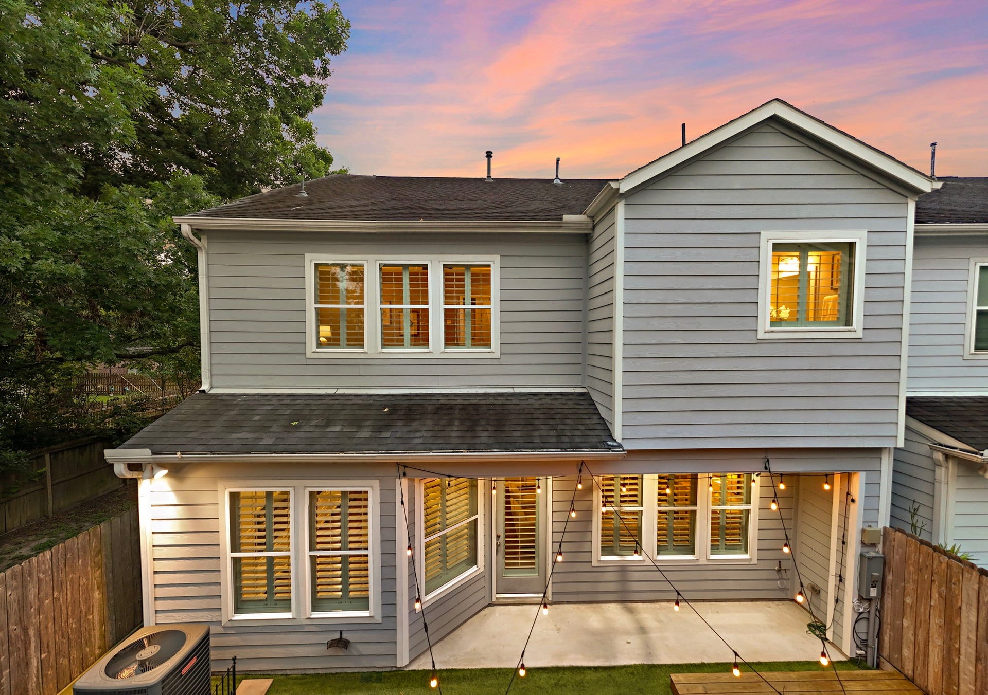4926 Ella Boulevard, Houston, TX 77018 Charming two-story home exterior with gray siding and large windows. The backyard features string lights over a cozy patio area, ideal for entertaining. Lush greenery and a wooden fence provide privacy, creating a serene outdoor space.