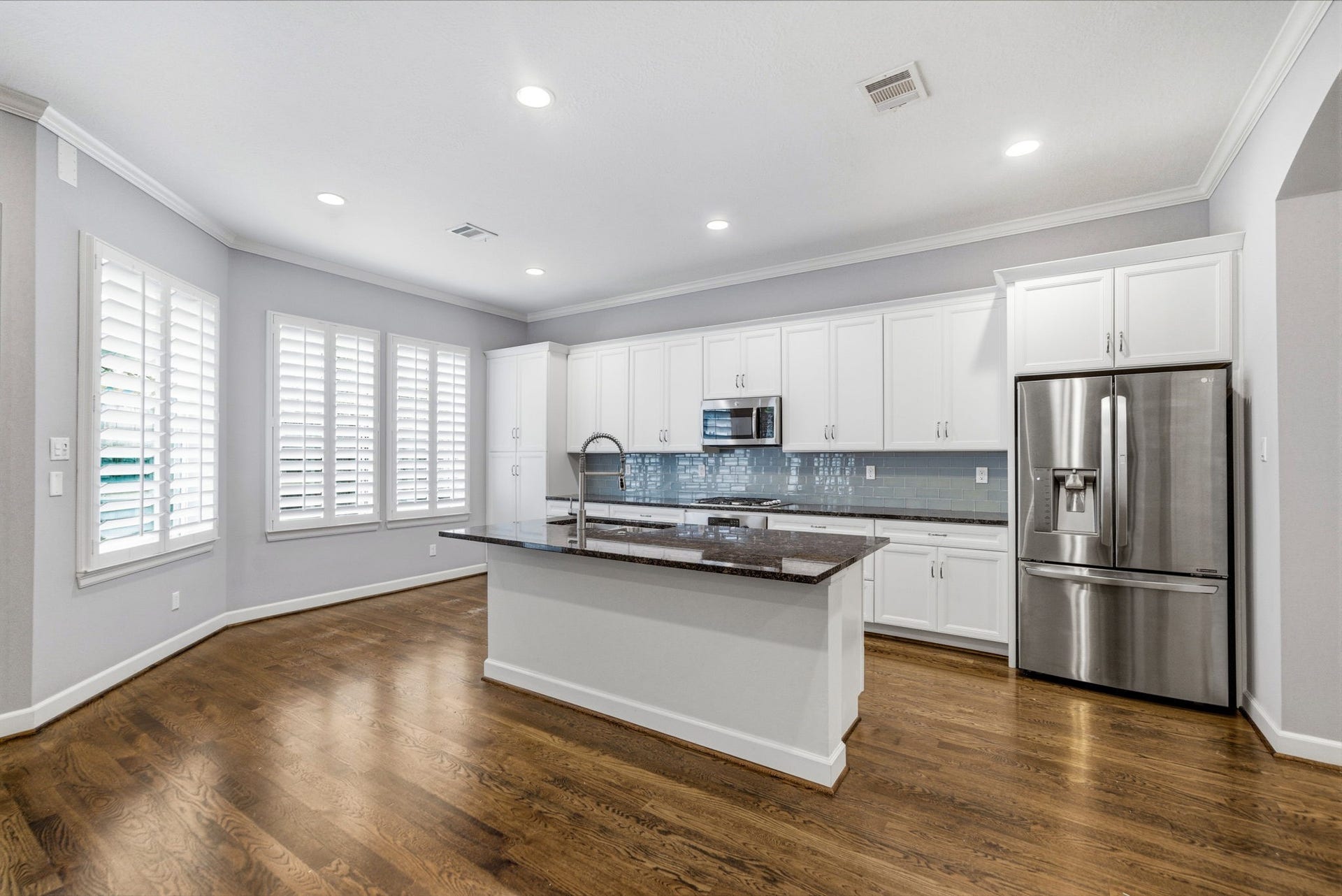 4926 Ella Boulevard, Houston, TX 77018 Bright and modern kitchen featuring hardwood floors, white cabinetry, stainless steel appliances including refrigerator and a central island. Large windows with shutters provide ample natural light.