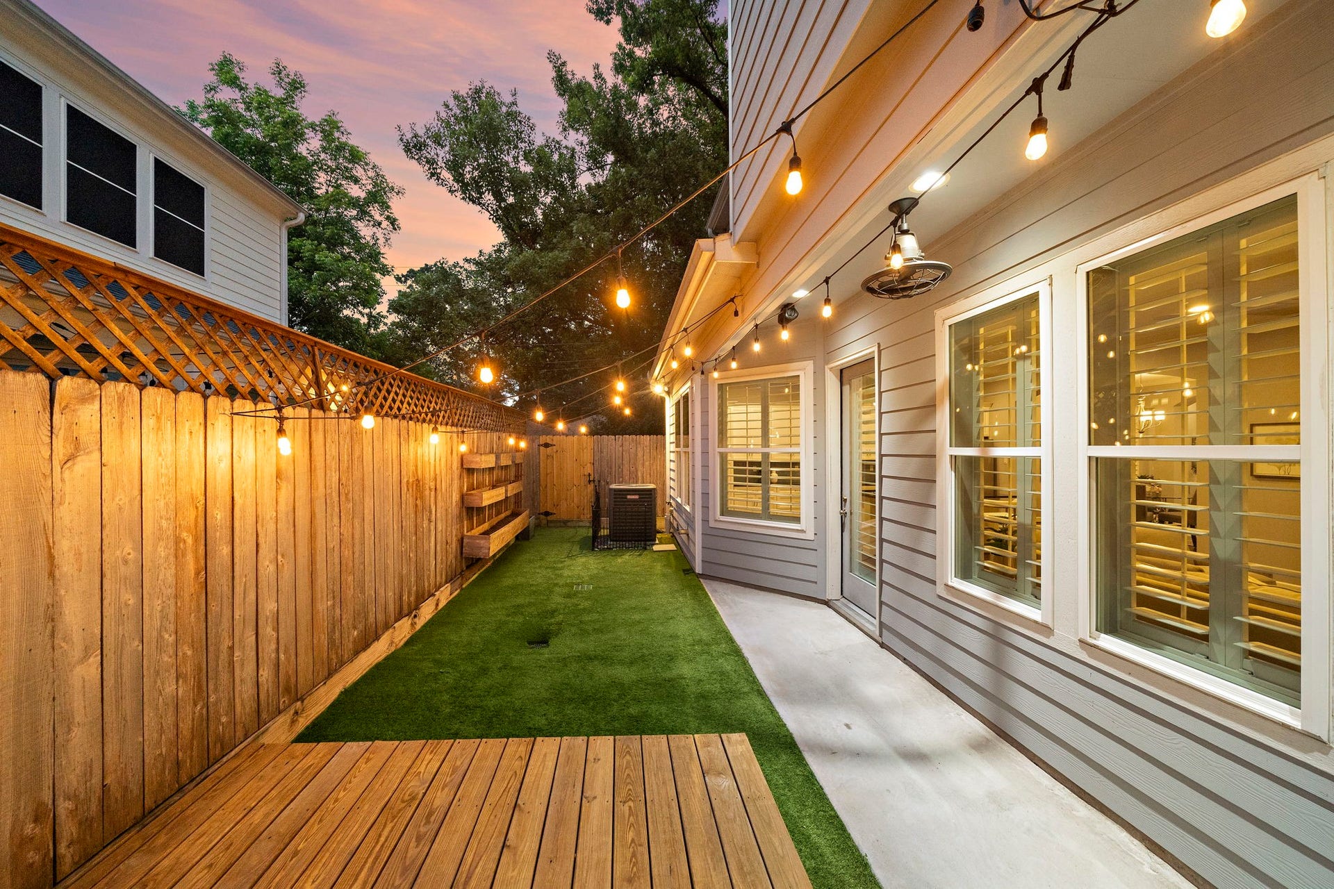 4926 Ella Boulevard, Houston, TX 77018 Charming backyard space featuring a wooden deck and artificial grass area. It is enclosed by a wooden fence adorned with string lights, creating a cozy and inviting atmosphere.