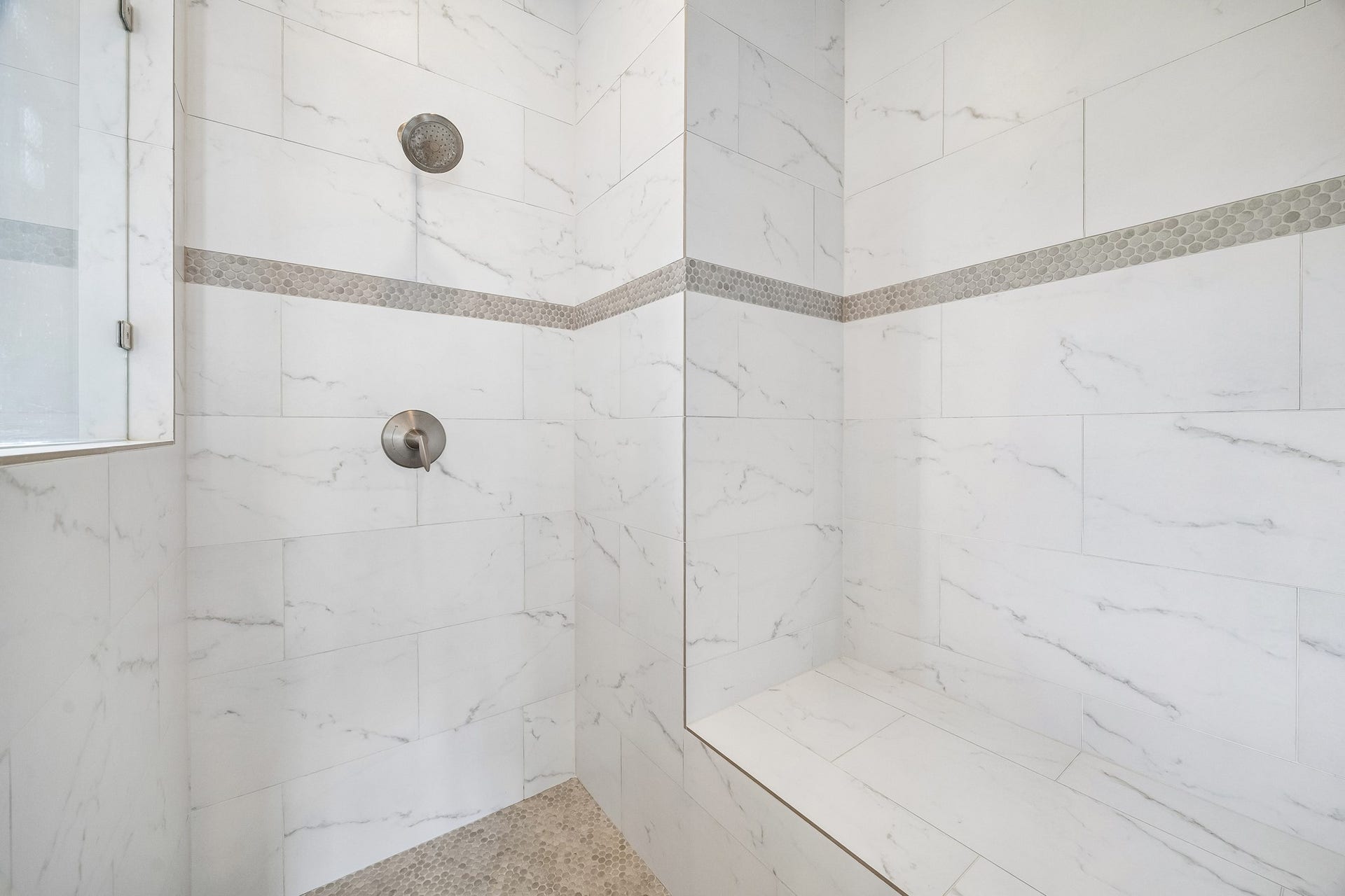 Inside view of the ample sized walk in shower updated in 2021 with sleek white marble tiles, accented by a decorative mosaic strip and a built-in bench for comfort.