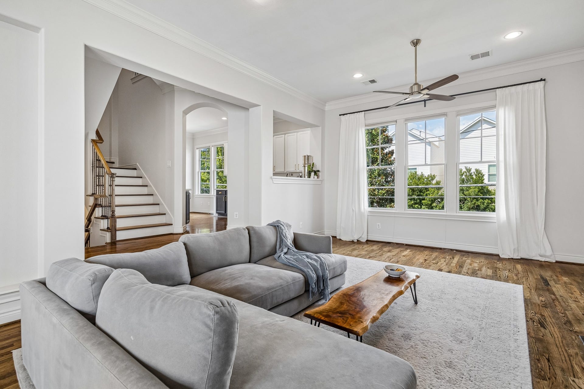 Bright and inviting living room featuring crown moldings, large windows, and elegant hardwood floors, with an open view to the kitchen and staircase.