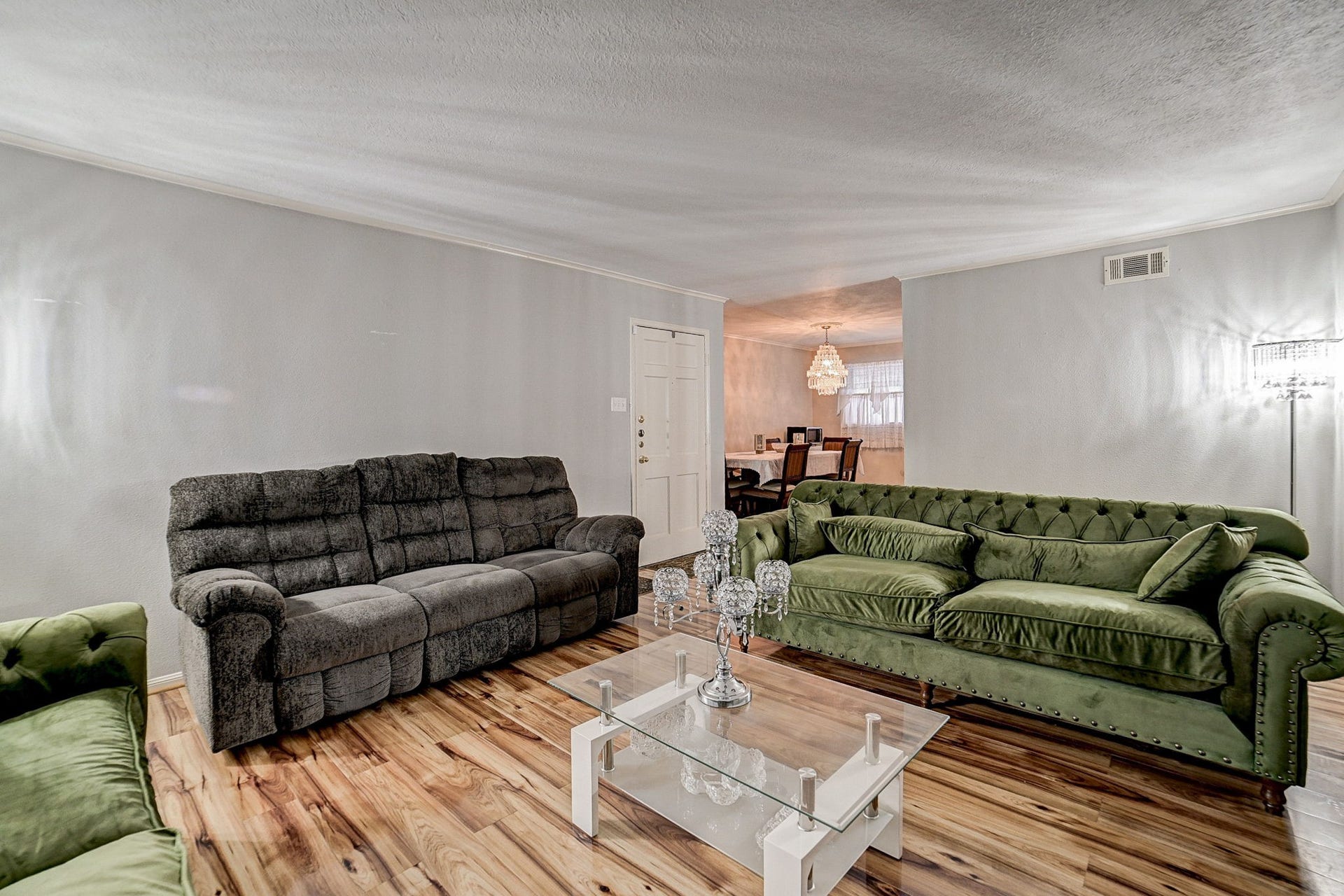 7900 Westheimer Road, #202, Houston, TX 77063 Spacious living room featuring elegant green and gray sofas, a chic glass coffee table, and warm wood flooring, leading into a cozy dining area illuminated by a stunning chandelier.
