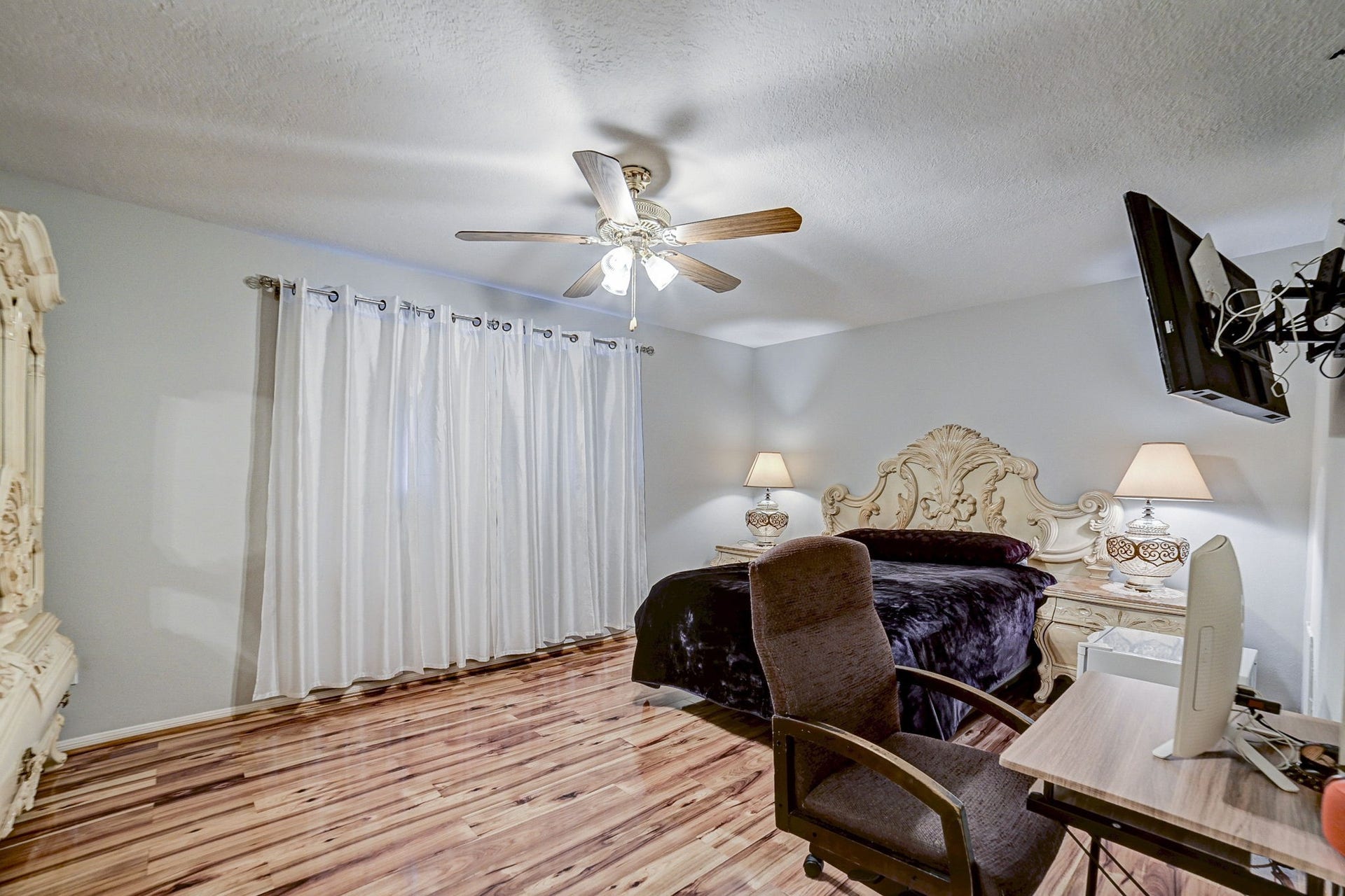 7900 Westheimer Road, #202, Houston, TX 77063 Spacious bedroom featuring elegant wood flooring, a stylish ceiling fan, and a luxurious bed with matching nightstands, complemented by soft lighting and a mounted TV for added convenience.