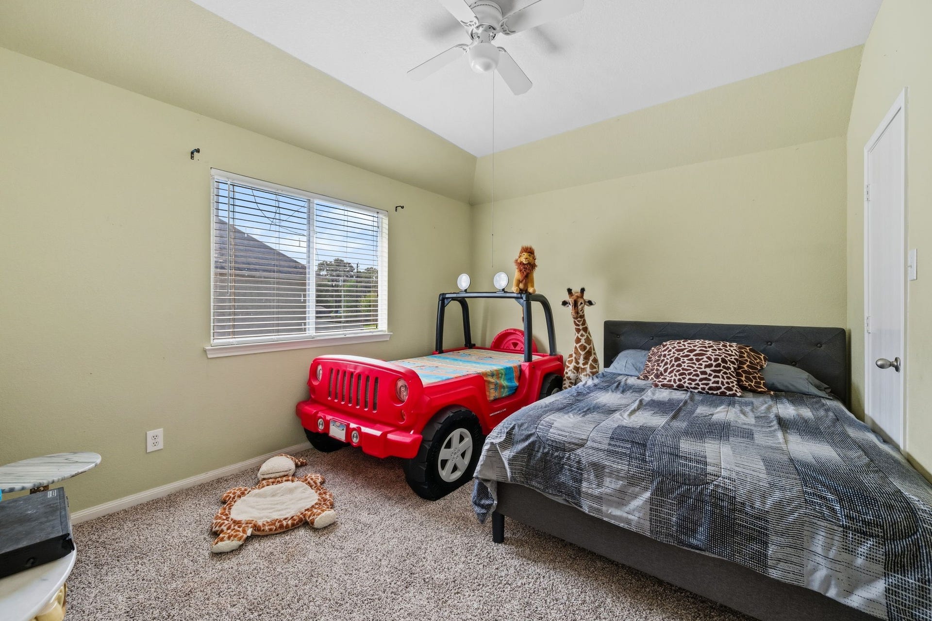 Charming children's bedroom featuring playful decor, including a red car-themed bed and plush animal accents, complemented by soft carpeting and natural light from a large window.