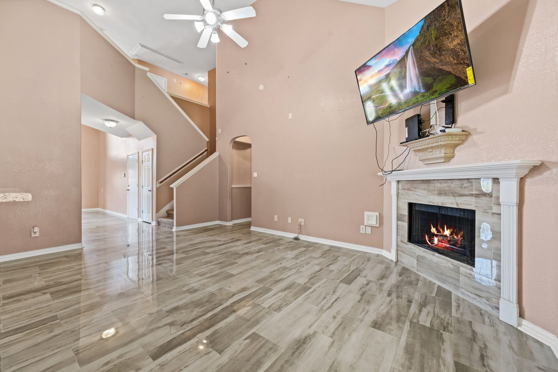 Spacious living area with high ceilings, elegant tile flooring, a cozy fireplace, and a mounted TV, complemented by a graceful staircase leading to the upper level.