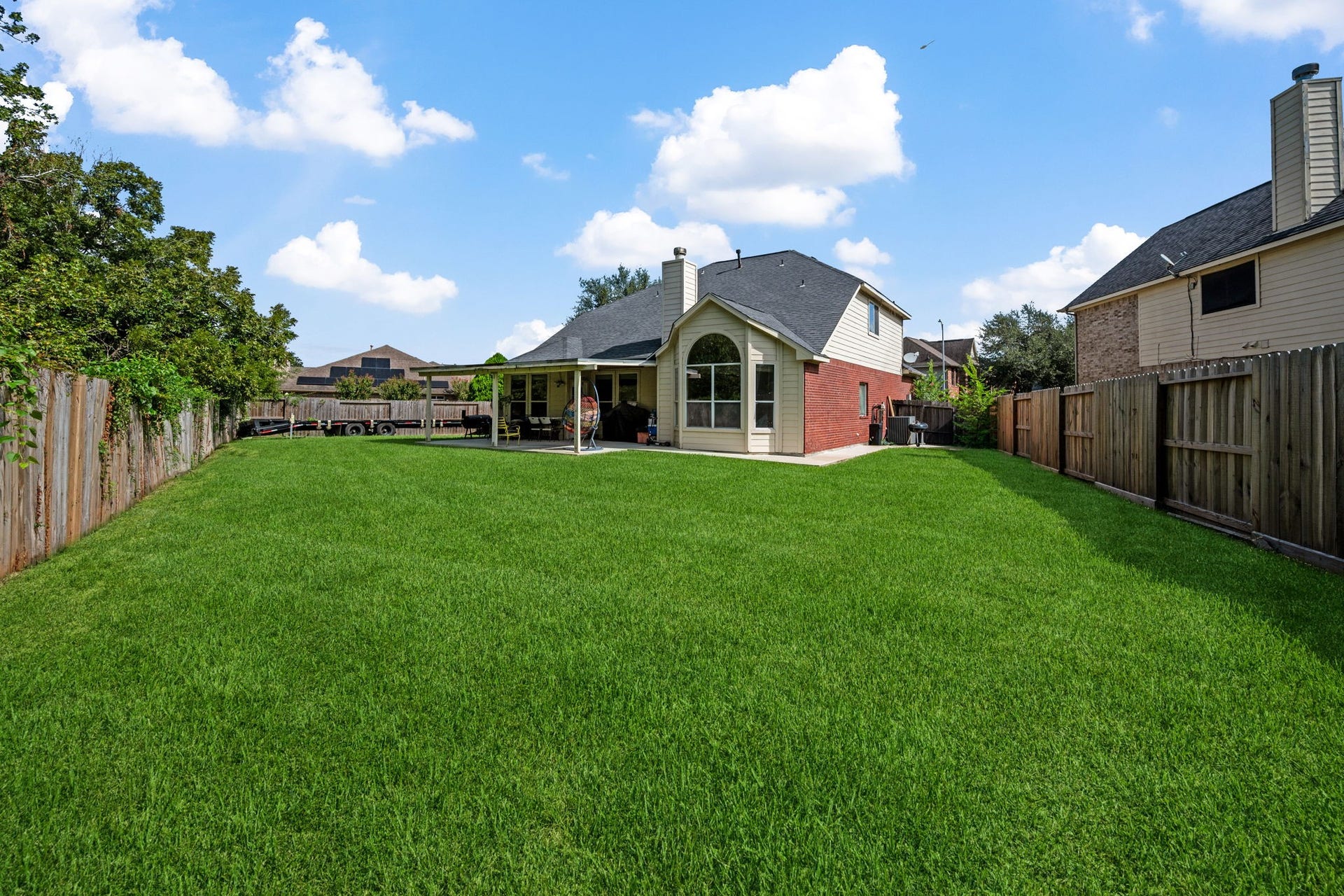 Expansive backyard featuring lush green grass, a charming brick house with large arched windows, and a cozy covered patio perfect for outdoor relaxation.