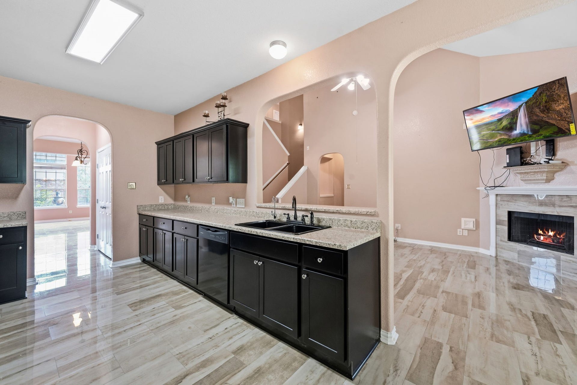 Elegant kitchen with sleek dark cabinetry, granite countertops, and polished tile flooring, seamlessly flowing into a cozy living area with a fireplace and mounted TV.
