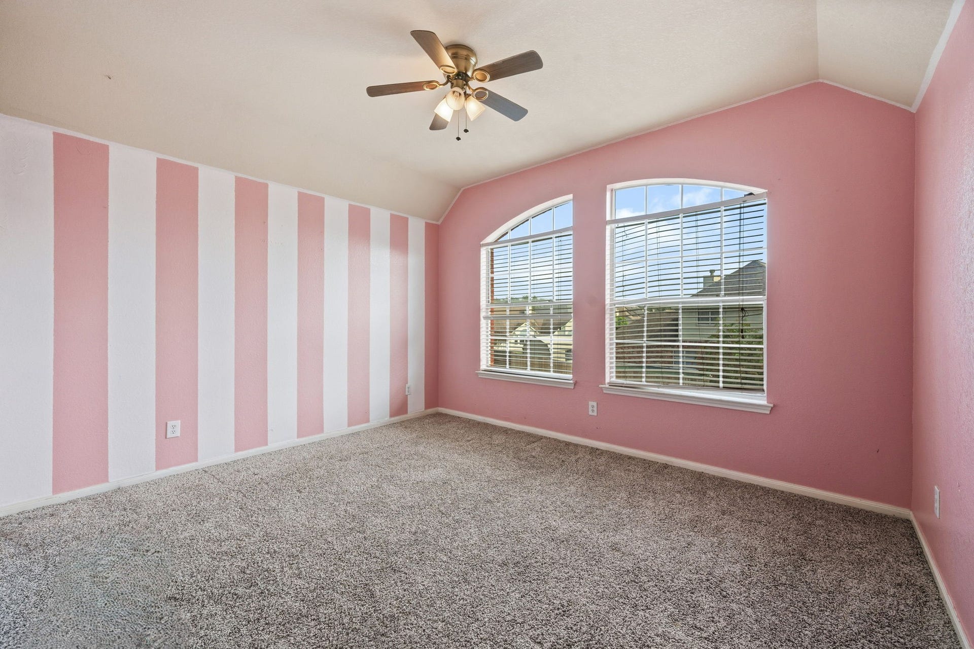 Bright and airy bedroom featuring charming pink and white striped walls, plush carpeting, and large arched windows that invite abundant natural light.
