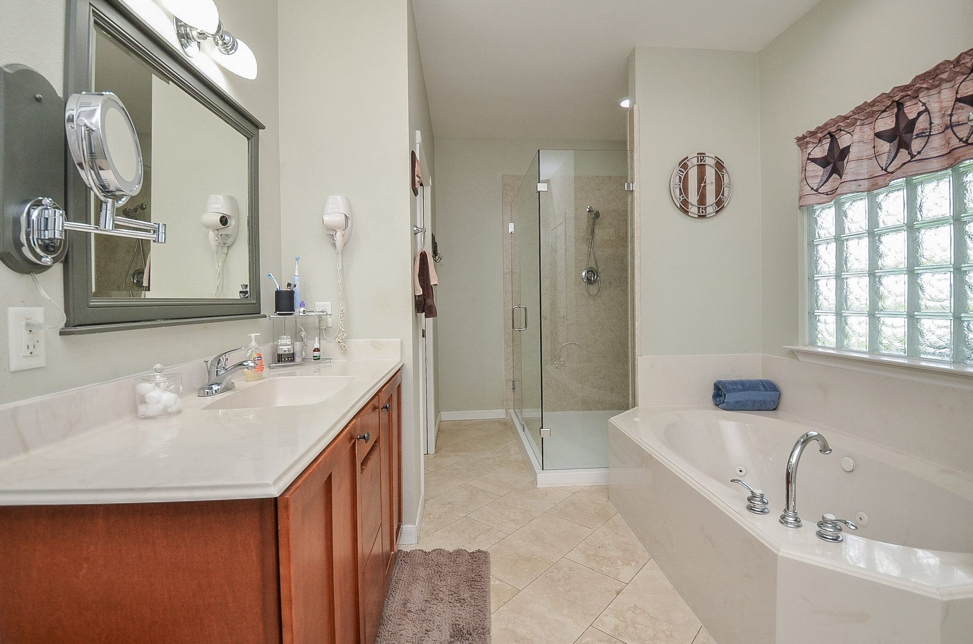 27644 Spring Hill Road, Hempstead, TX 77445 Clean, streamlined and comfortable paths line your primary ensuite bath; with lush honey colored cabinetry (left); pale earth toned walls, quaint art deco-like window above the generous soaking tub; with clear glass enclosed shower, up right.