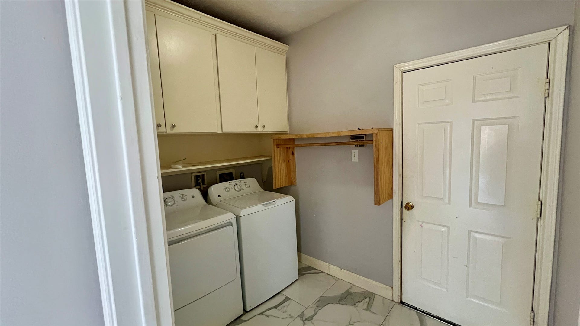 This photo shows a laundry room with a washer and dryer, white cabinetry for storage, and a door leading outside
