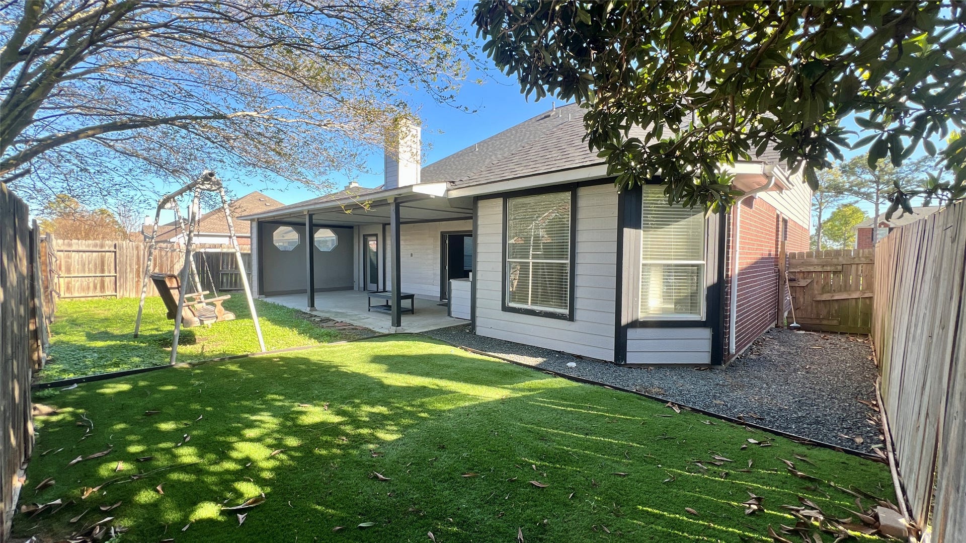 Charming backyard featuring a covered patio, swing set, and artificial turf for easy maintenance. Surrounded by a wooden fence, it offers privacy and a cozy outdoor space.