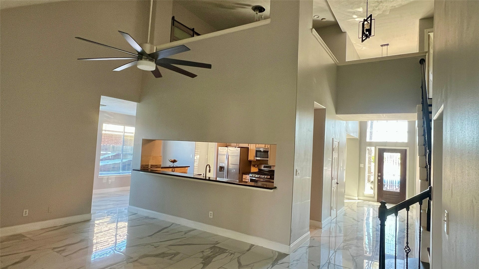 This photo showcases a spacious, modern interior with high ceilings, a large ceiling fan, and glossy marble-like flooring. The open layout features a view of the kitchen through a passthrough window and a grand entrance with ample natural light.