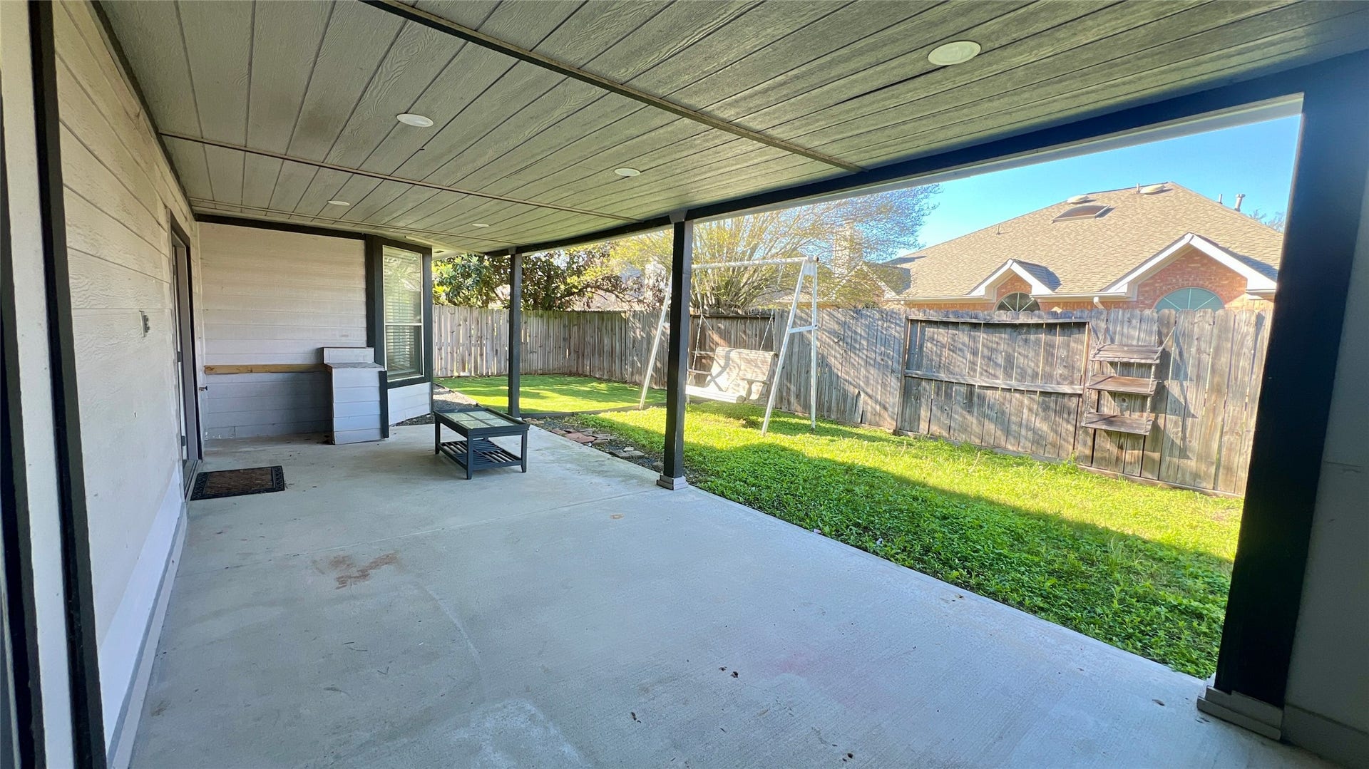 Covered patio area with a concrete floor, overlooking a grassy backyard. The space is enclosed by a wooden fence, offering privacy and potential for outdoor activities or relaxation.