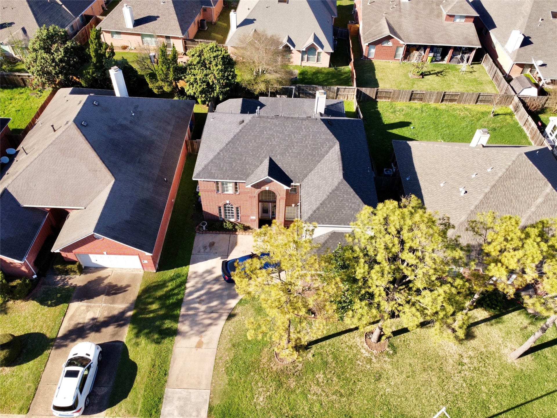 This aerial view showcases a two-story brick home with a spacious driveway and a well-maintained front lawn. It is situated in a suburban neighborhood with similar houses, offering a fenced backyard and mature trees for added privacy.