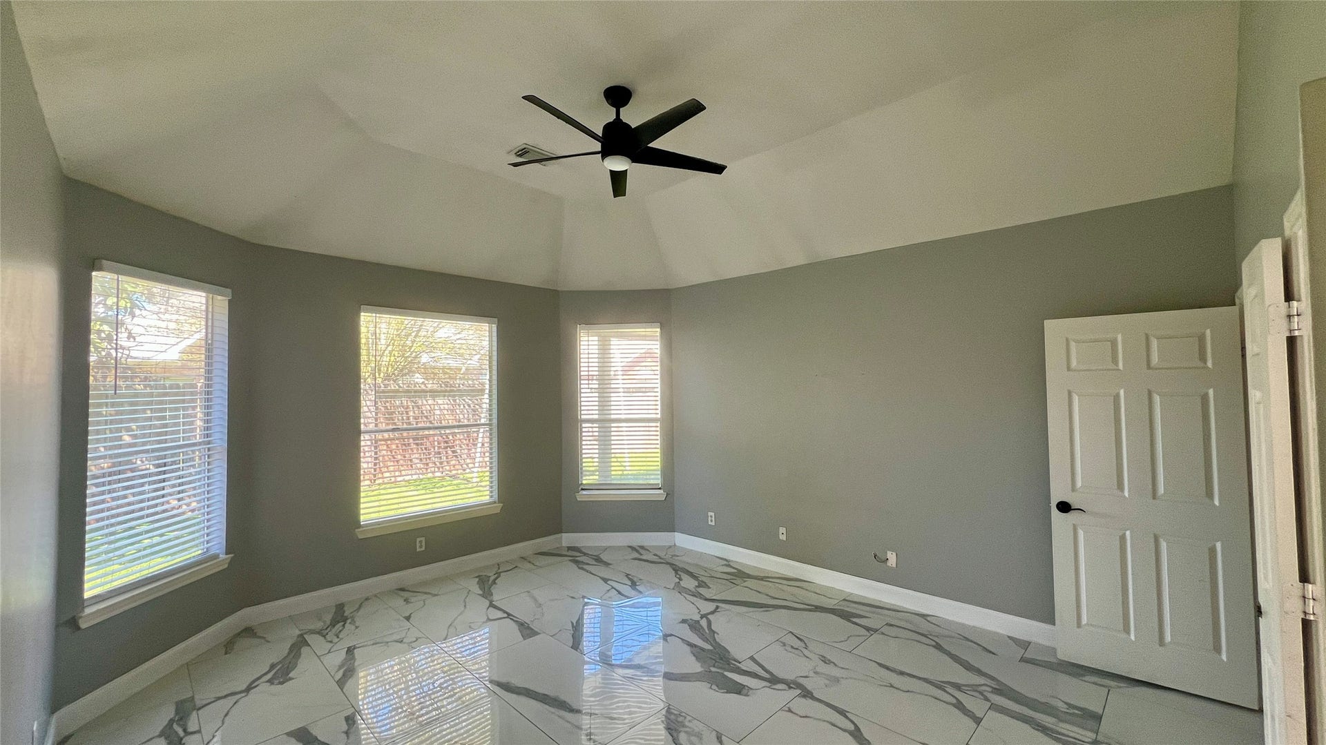 This room features large windows offering plenty of natural light, neutral gray walls, glossy marble-patterned tile flooring, and a modern ceiling fan.