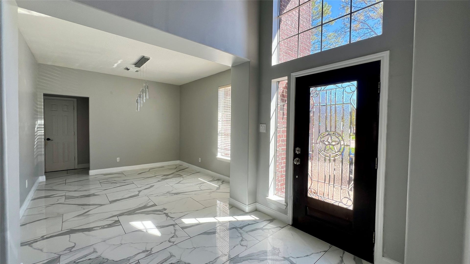This bright entryway features large windows, a decorative front door, and sleek marble-tiled flooring. The adjacent room offers modern lighting and ample natural light, creating an inviting and stylish atmosphere