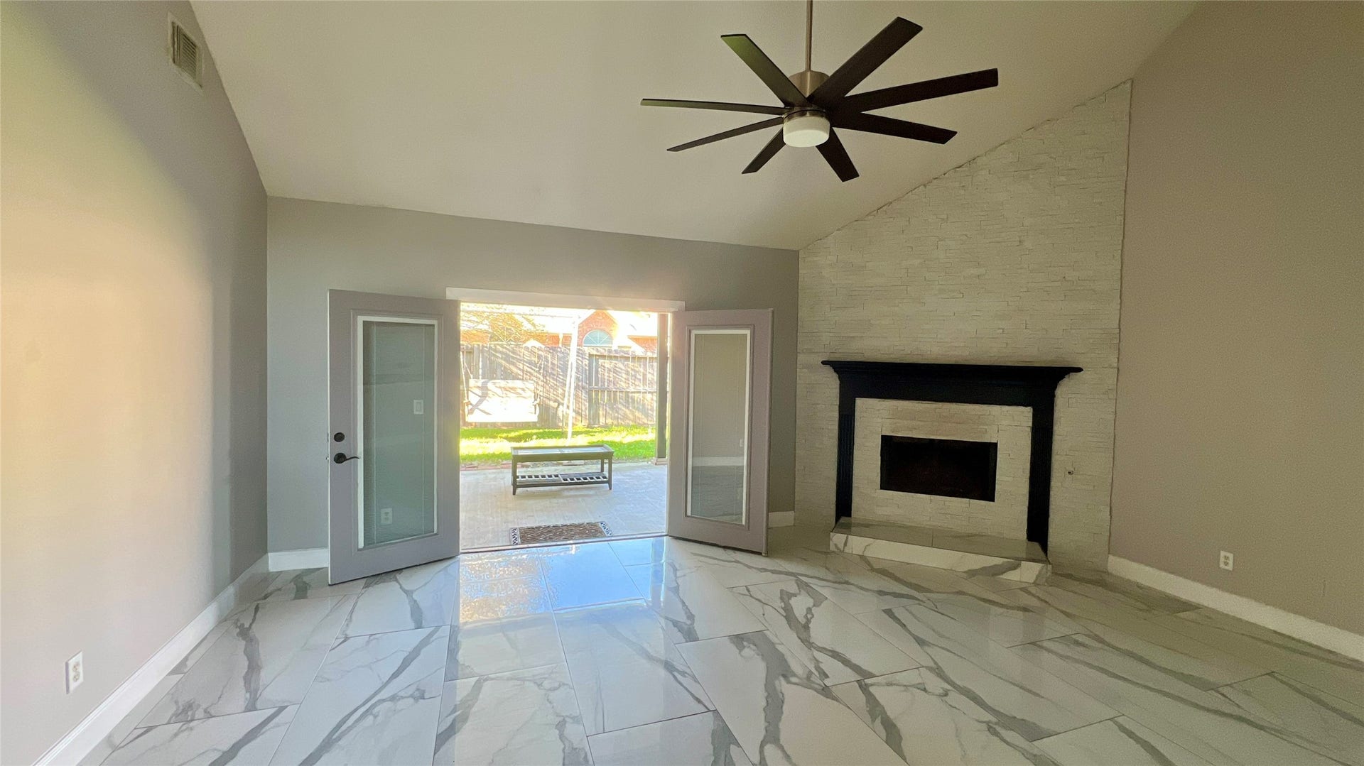 This spacious living area features high ceilings, a modern ceiling fan, and a stylish fireplace with a dark mantel. It boasts large, elegant marble-like tiles and French doors that lead to a backyard, offering plenty of natural light and indoor-outdoor flow.