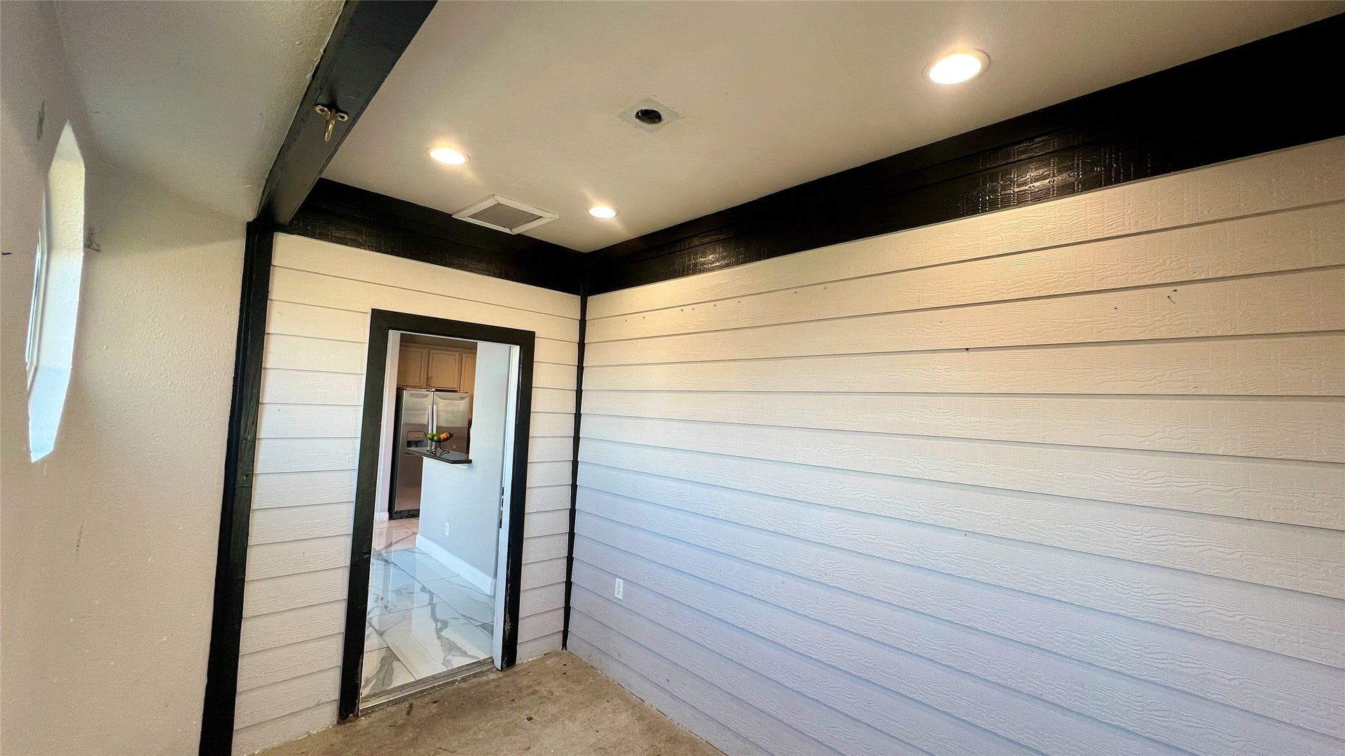 This photo shows a narrow room with white panel walls and a dark trim. Recessed lighting brightens the space, and there's an open doorway leading to a kitchen area