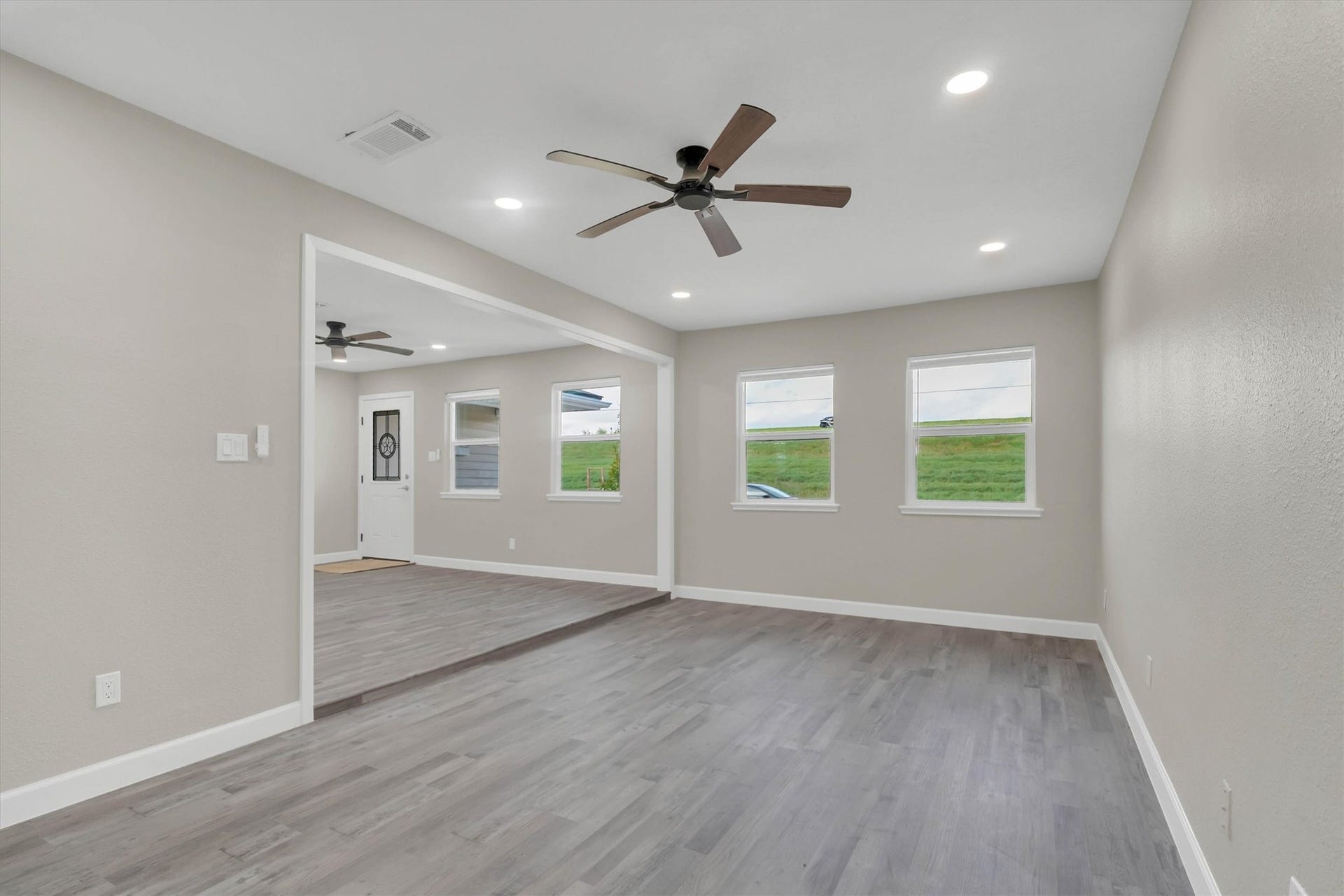 Spacious living area with modern gray flooring, recessed lighting, and ceiling fans, accented by large windows for natural light.