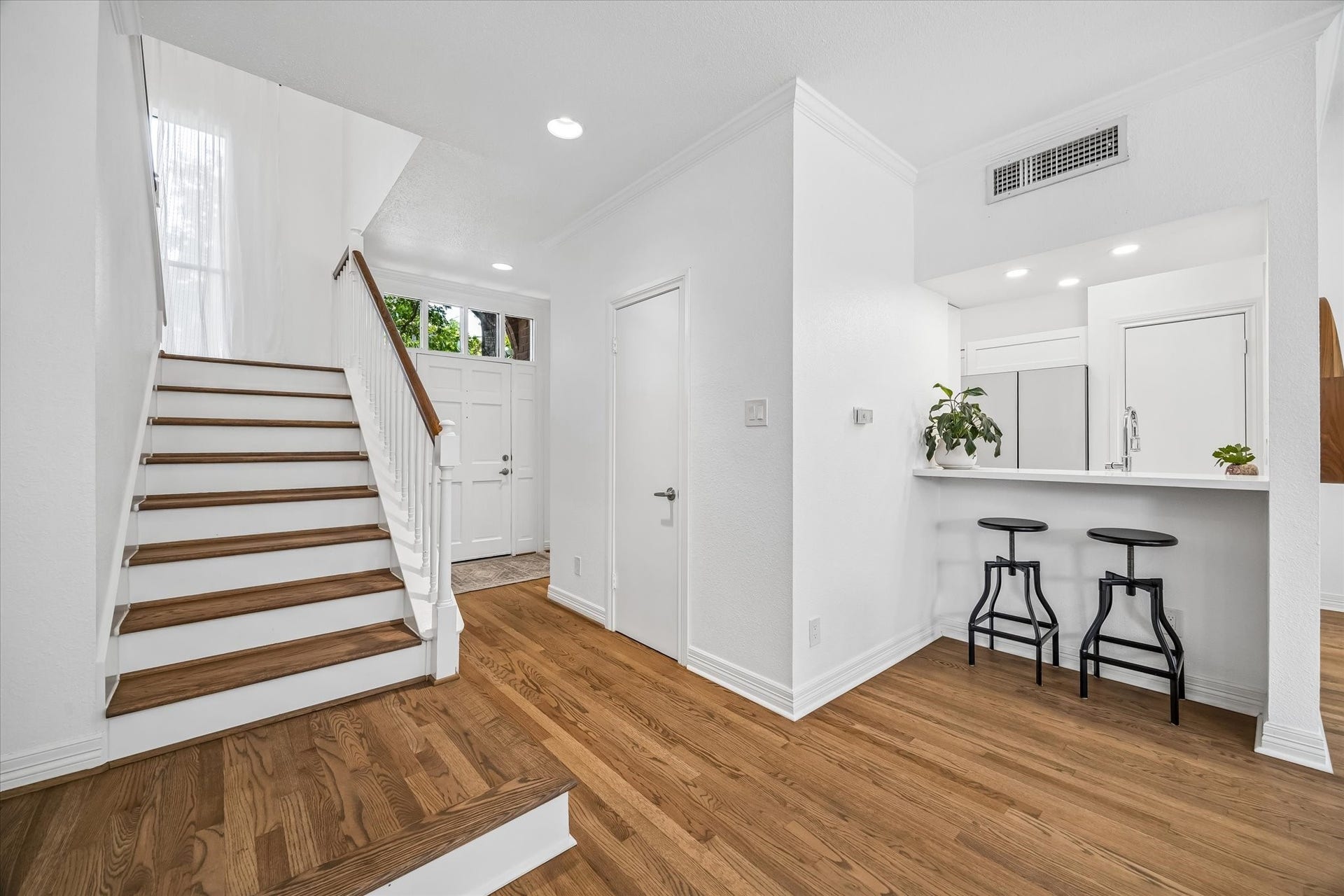 Bright and inviting entryway featuring elegant hardwood floors, a modern staircase, and a cozy breakfast bar leading to a well-lit kitchen area.