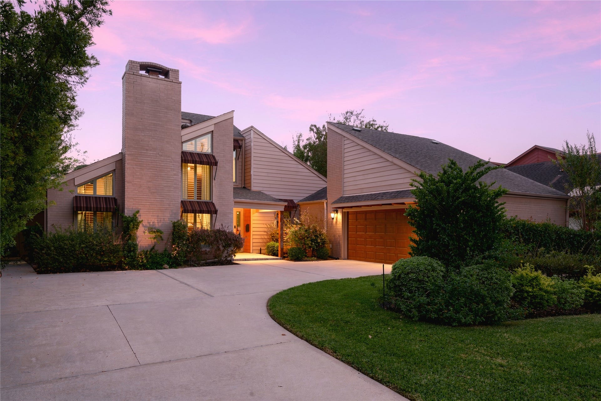 3211 Chris Drive, Houston, TX 77063 Striking contemporary exterior featuring a tall brick chimney, striped metal awnings & glowing windows at dusk, set behind a sweeping curved driveway & manicured lawn.
