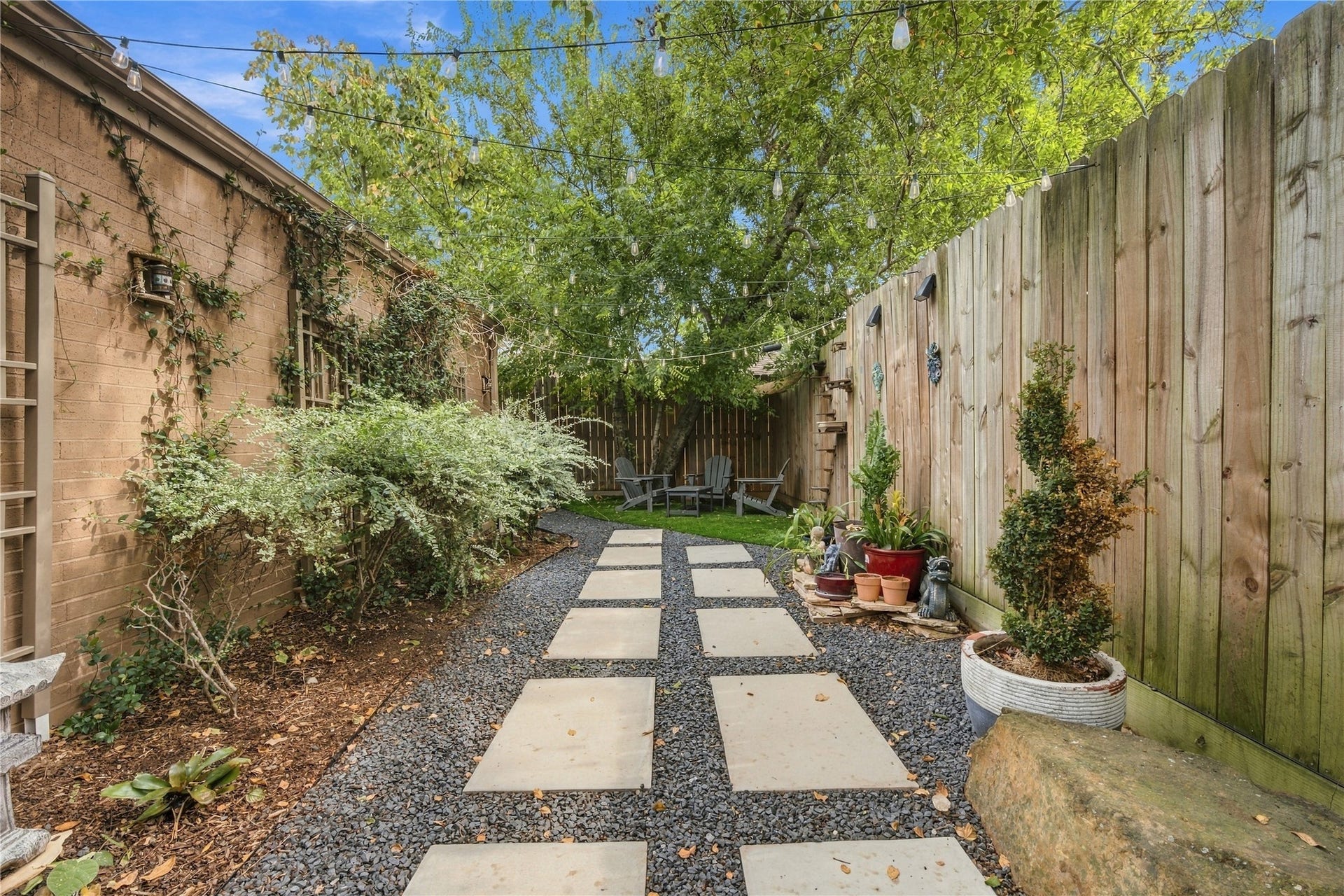 3211 Chris Drive, Houston, TX 77063 Square stepping stones set in gravel lead down the side yard beneath a canopy of string lights, between a tall wood privacy fence & planted beds along the brick. The path ends at a turf seating area under shaded trees.
