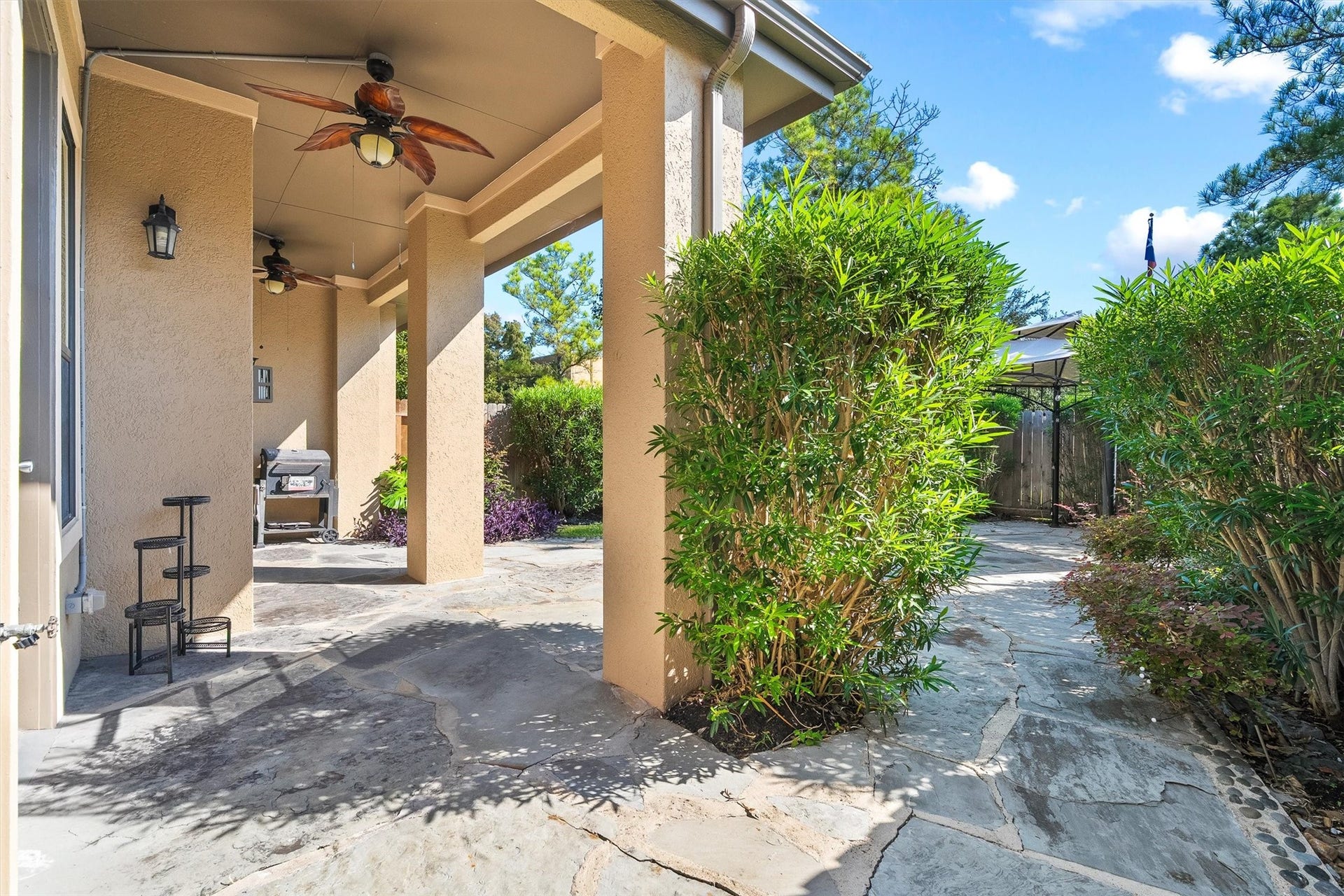 2919 Royal Oaks Crest, Houston, TX 77082 A covered patio with ceiling fans and ample space for outdoor entertaining, surrounded by lush greenery and a stone pathway leading to a cozy seating area.