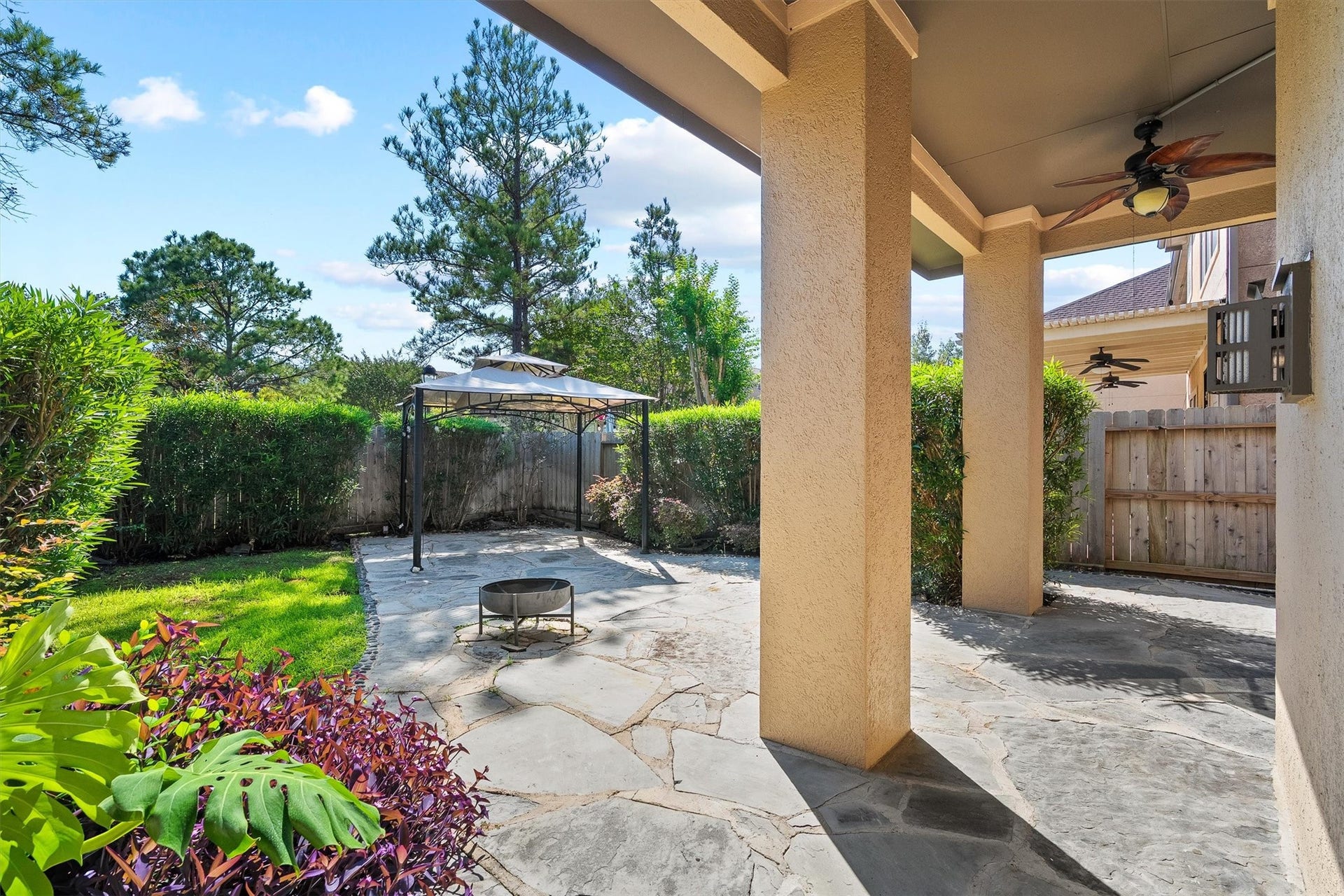 2919 Royal Oaks Crest, Houston, TX 77082 A backyard featuring a covered patio with ceiling fans, a stone-paved area with a fire pit, and a gazebo, all surrounded by lush greenery for privacy and relaxation.