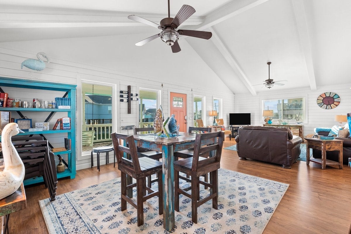 2406 Avenue P, San Leon, TX 77539 This inviting open-concept living space features high vaulted ceilings, abundant natural light, and stylish wood flooring. The room is tastefully decorated with coastal accents, including a rustic dining set and cozy seating area. Large windows and a charming front door enhance the airy, welcoming atmosphere.