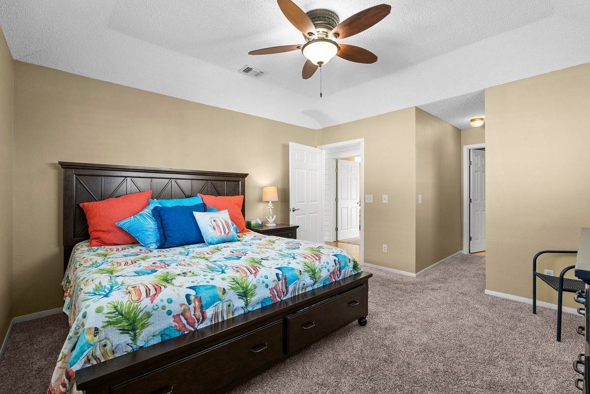 2406 Avenue P, San Leon, TX 77539 Spacious bedroom with a cozy carpet, featuring a large bed with colorful bedding. The room includes a ceiling fan, neutral walls, and ample natural light, providing a warm and inviting atmosphere.