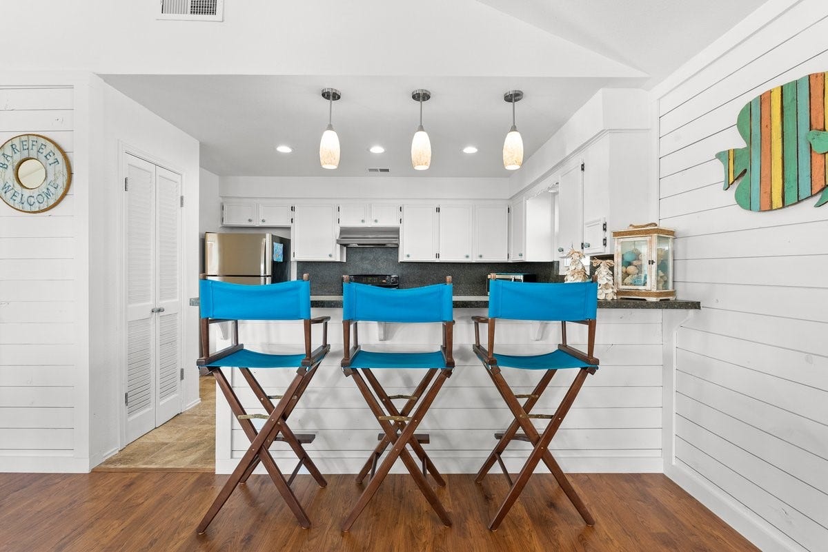 2406 Avenue P, San Leon, TX 77539 Bright, coastal-style kitchen with white cabinets and shiplap walls. Features a breakfast bar with three blue director's chairs, pendant lighting, and stainless steel appliances.