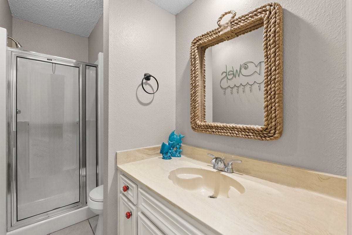 2406 Avenue P, San Leon, TX 77539 This bathroom features a clean, modern design with a glass-enclosed shower, a beige countertop with a sink, and a decorative rope-framed mirror. The neutral tones and simple decor create a welcoming, functional space.