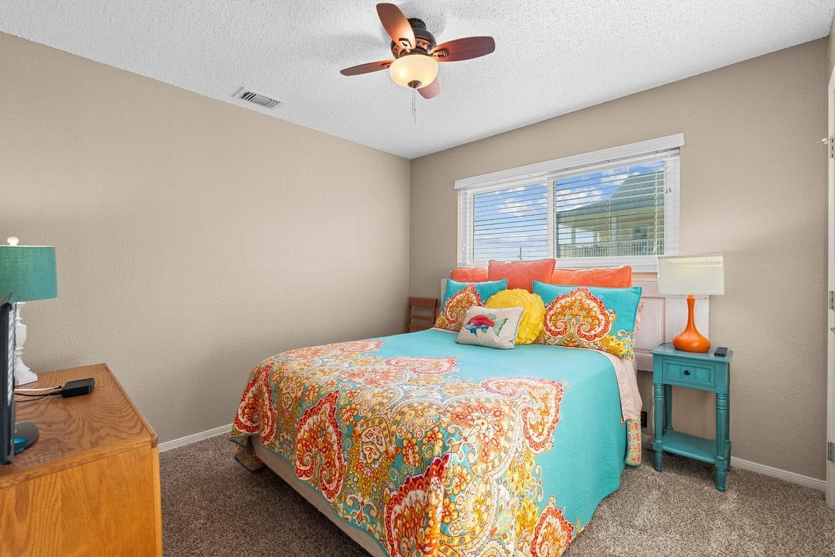 2406 Avenue P, San Leon, TX 77539 Cozy bedroom with neutral walls and carpet, featuring a colorful bedspread and pillows. It has a ceiling fan, a large window for natural light, and a mix of wooden and painted furniture for a vibrant touch.