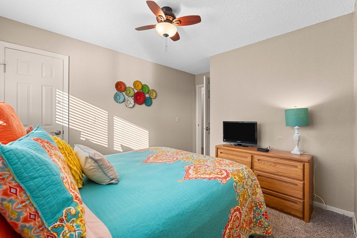 2406 Avenue P, San Leon, TX 77539 This cozy bedroom features warm beige walls and a colorful bedspread, creating a vibrant, inviting atmosphere. The room includes a ceiling fan offering both comfort and style.