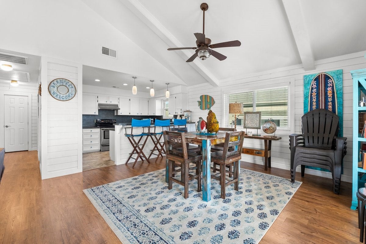 2406 Avenue P, San Leon, TX 77539 This bright, open-concept living space features a vaulted ceiling and hardwood floors. The kitchen has white cabinets, stainless steel appliances, and a breakfast bar with blue stools. The dining area is cozy, with colorful decor creating a welcoming atmosphere.
