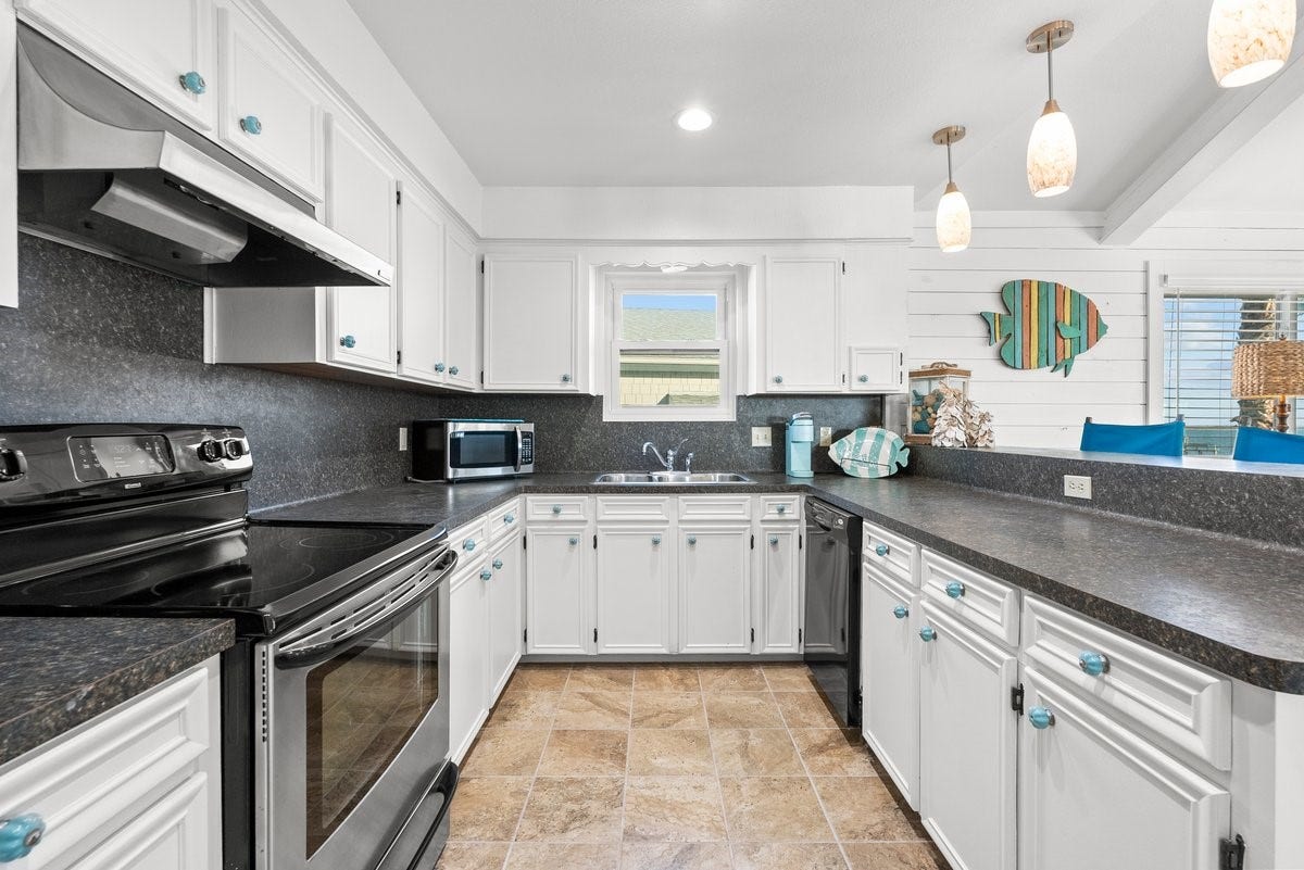 2406 Avenue P, San Leon, TX 77539 This kitchen features a bright, coastal design with white cabinetry, dark countertops, and stainless steel appliances. It includes a spacious layout with ample counter space, modern light fixtures, and decorative accents, creating a welcoming and functional cooking area.