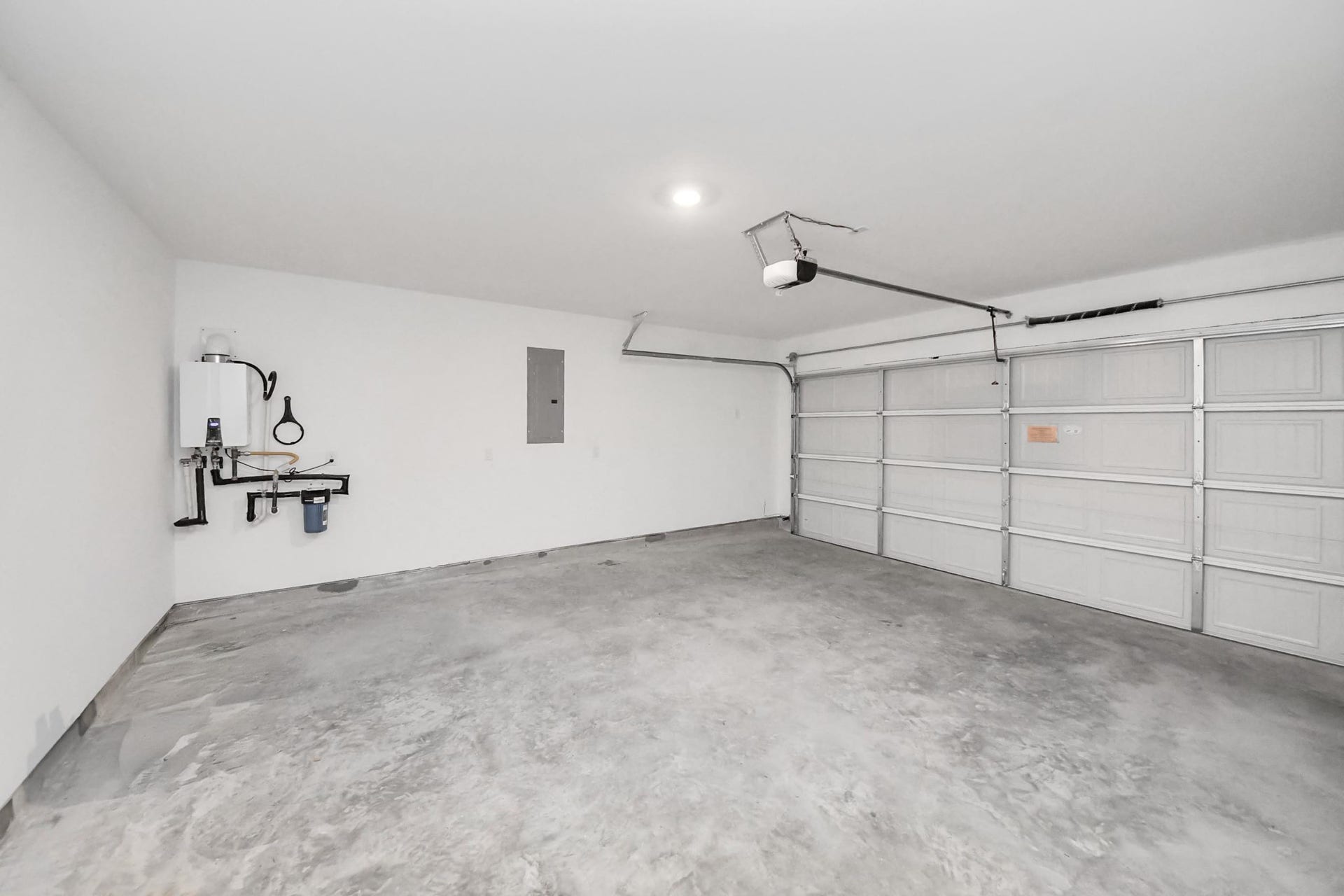 3096 Sunset Glory Drive, Katy, TX 77493 This photo shows a spacious, clean garage with a concrete floor and white walls. It features a large double garage door with an automatic opener and a wall-mounted water heater. Ideal for parking and storage.