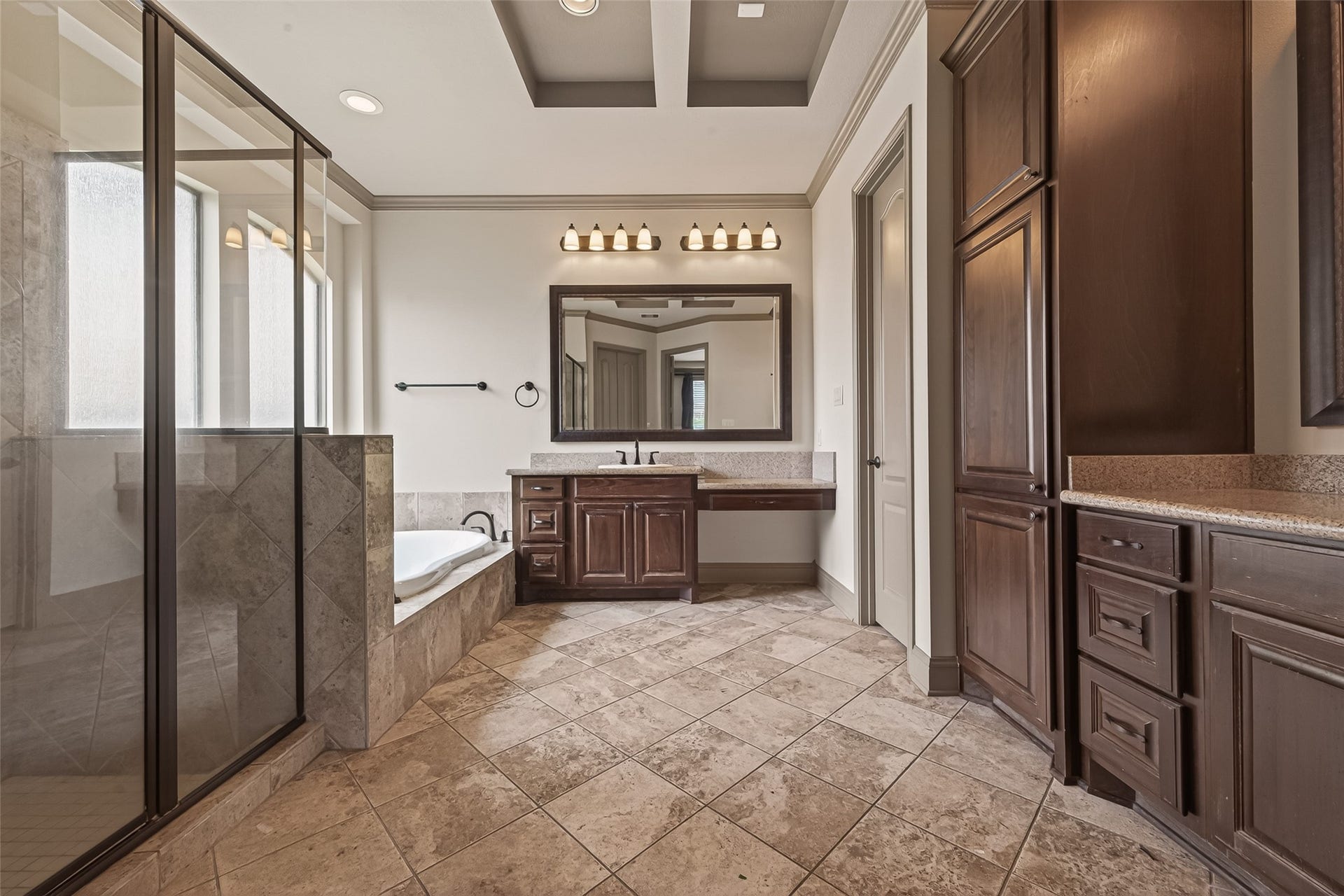 27411 Manor Falls Lane, Fulshear, TX 77441 The spacious primary bathroom features a tray ceiling with crown molding, tile flooring, and rich wood cabinetry with granite countertops. A glass enclosed shower and a soaking tub provide the perfect blend of luxury and practicality. A tall linen cabinet on the right offers generous storage, keeping the space organized and clutter free.