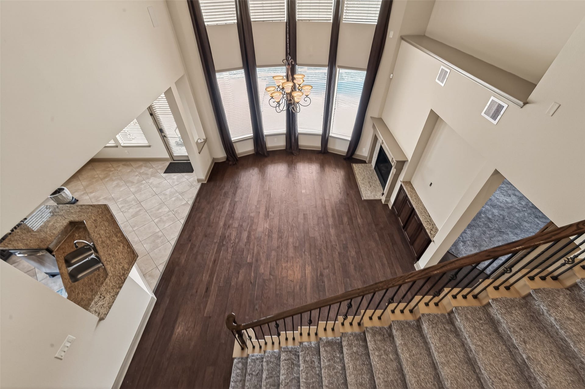 27411 Manor Falls Lane, Fulshear, TX 77441 From the upper level, this breathtaking view captures the grandeur of the home's open layout. Soaring two-story ceilings, a dramatic curved wall of windows, and an elegant chandelier create a striking focal point in the living area below. Rich hardwood flooring anchors the space, while a fireplace with a granite surround adds warmth and sophistication. The staircase with its wooden handrail and wrought iron balusters leads gracefully to this impressive vantage point, with the kitchen visible just beyond.