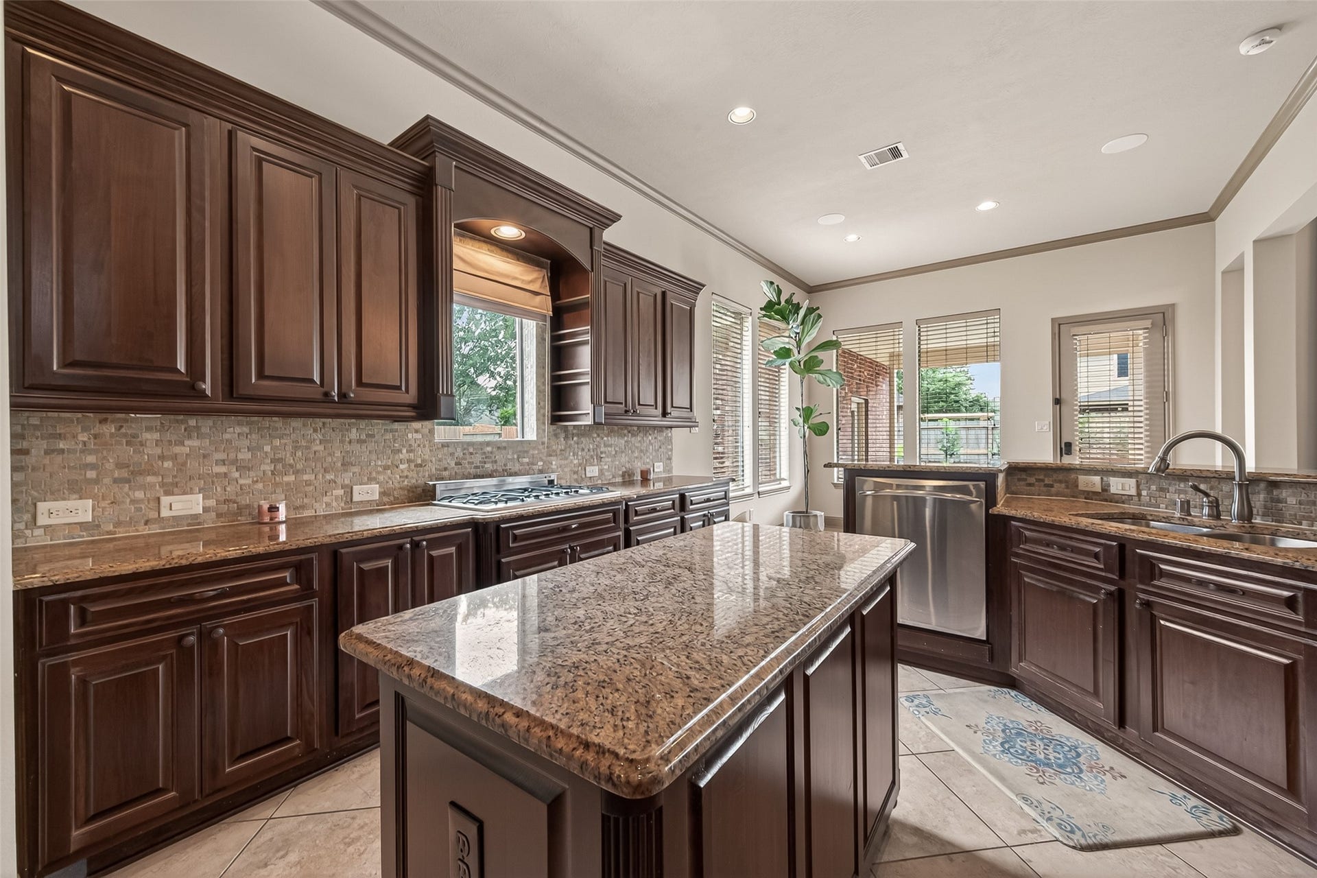 27411 Manor Falls Lane, Fulshear, TX 77441 From this angle, the kitchen's thoughtful layout truly shines. Rich dark wood cabinetry lines the walls, complemented by granite countertops and a stone mosaic backsplash. The gas cooktop is perfectly positioned beneath a window that fills the space with natural light, while multiple windows along the back wall keep the kitchen bright and airy. The central island offers generous prep space, and the stainless steel dishwasher adds everyday convenience. A dual-sink setup ensures effortless meal prep and clean