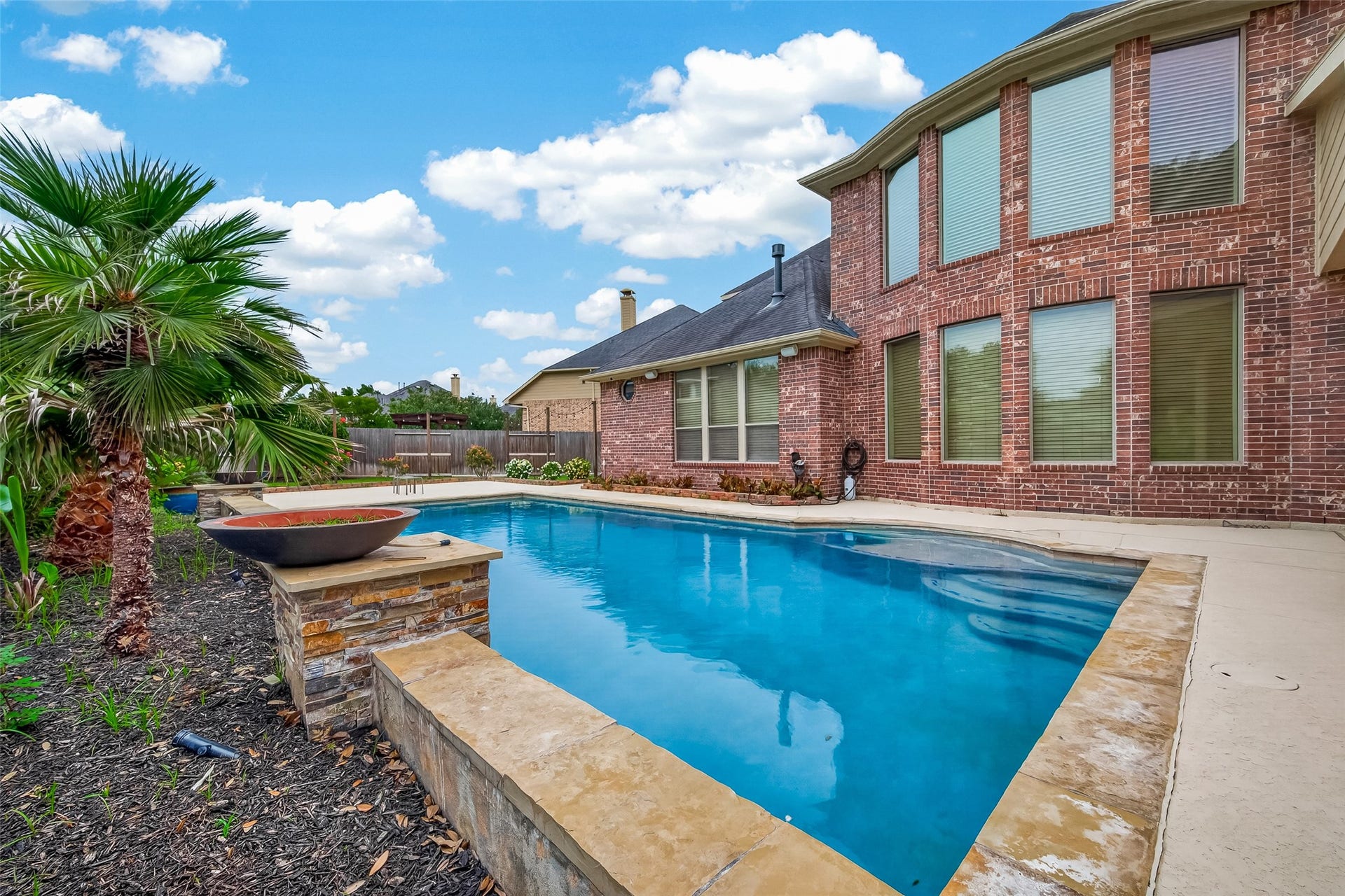 27411 Manor Falls Lane, Fulshear, TX 77441 This view from poolside showcases the stunning combination of the sparkling pool and the home's impressive brick rear elevation. A raised stone planter adds architectural interest to the pool area, while lush tropical palms and flowering plants create a vibrant, private backdrop. The covered patio just off the home provides a shaded retreat, making this backyard a perfect setting for entertaining loved ones or simply unwinding on a beautiful day.