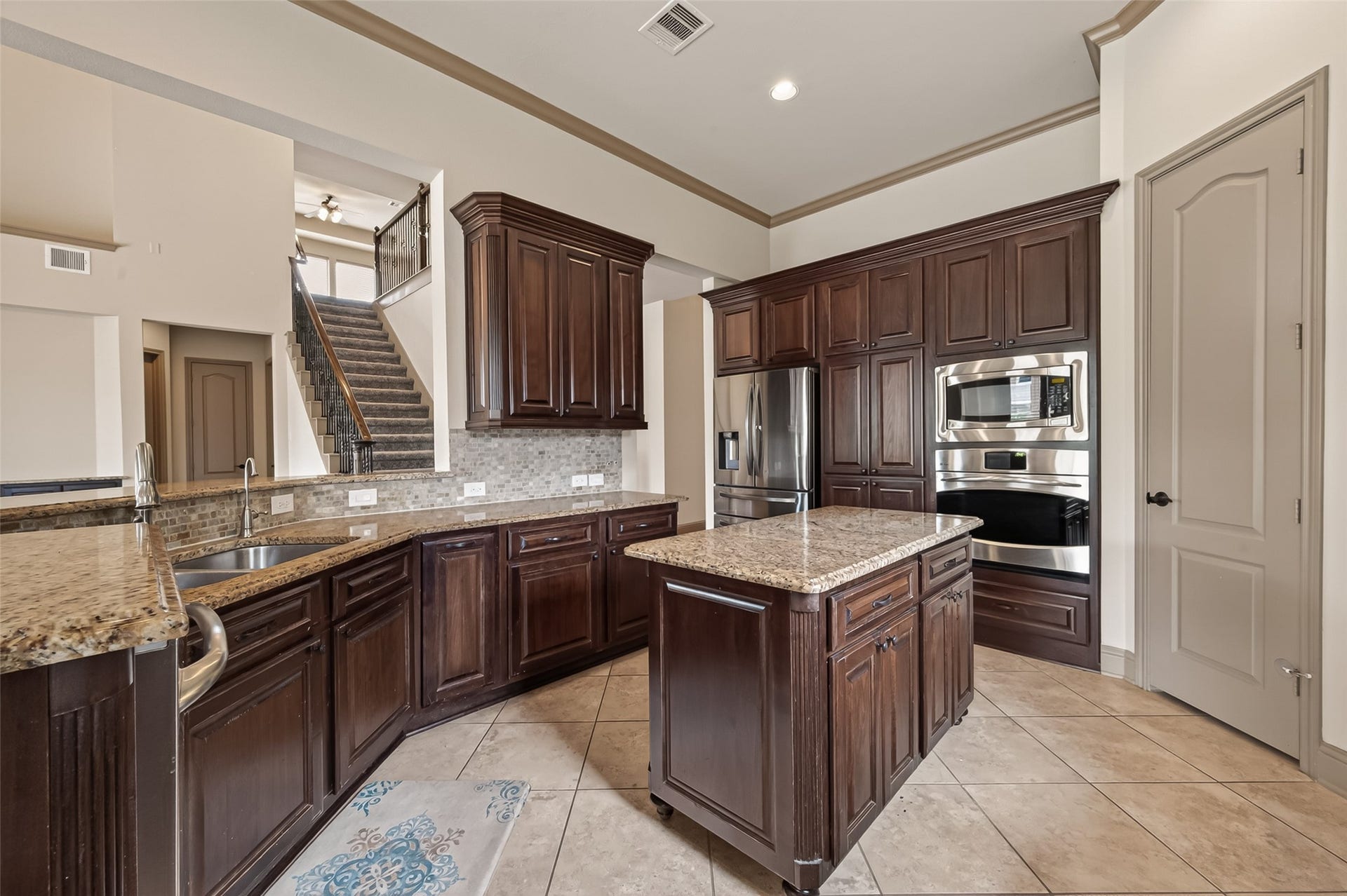 27411 Manor Falls Lane, Fulshear, TX 77441 This expansive gourmet kitchen is a chef's dream, featuring rich dark wood cabinetry with raised panel detailing, granite countertops, and a tasteful mosaic tile backsplash. Stainless steel appliances include a built-in double oven, microwave, and refrigerator with water dispenser. A central island provides additional prep space and storage, while tile flooring and crown molding add a refined finishing touch. From this vantage point, the elegant staircase with wrought iron railing is visible, showcasing the