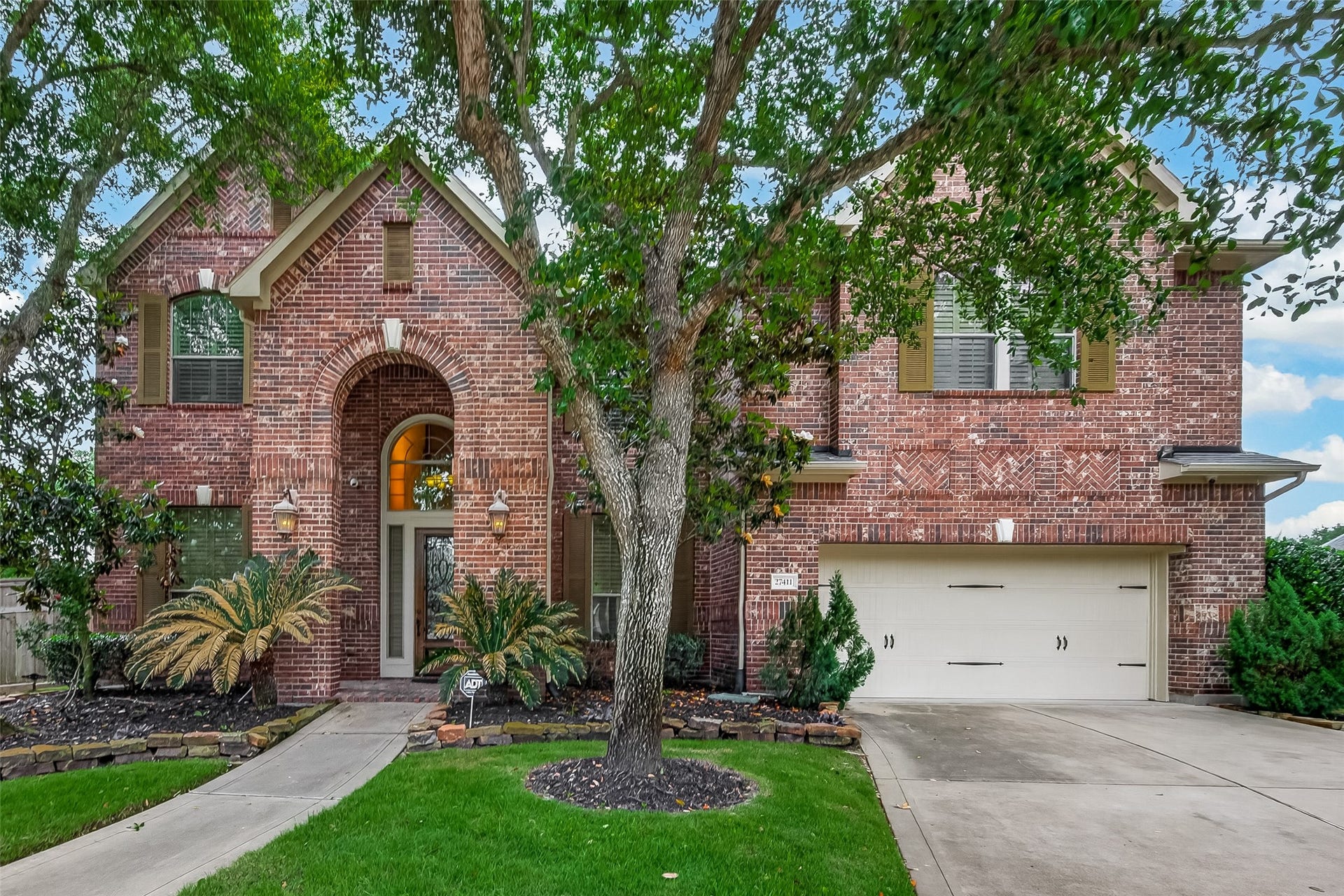 27411 Manor Falls Lane, Fulshear, TX 77441 This stunning two-story brick home makes a lasting first impression with its classic red brick facade, decorative herringbone brickwork accents, and a beautiful arched entryway framed by coach lantern lighting. A mature tree anchors the front lawn, adding natural shade and character to the lush, well-maintained landscaping. The two-car attached garage and wide driveway provide ample parking, while the thoughtfully designed plant beds with tropical foliage complete this home's gorgeous curb appeal.