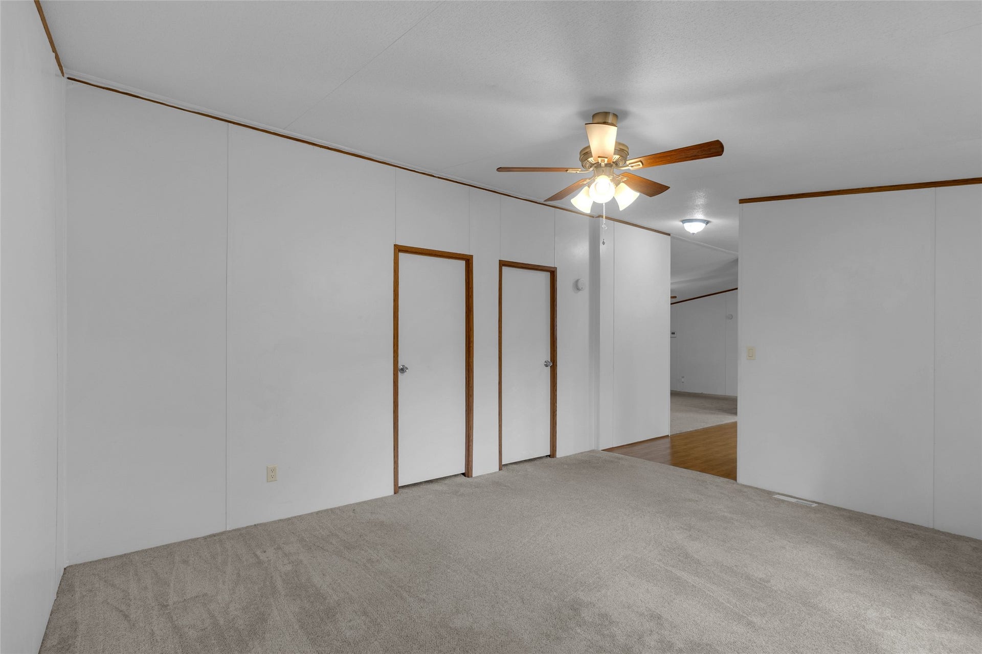 Den room features neutral-toned carpeting, white walls with wood trim, a ceiling fan, and recessed lighting, offering a simple and functional space. The area includes two doors and a transition into the two additional bedrooms.