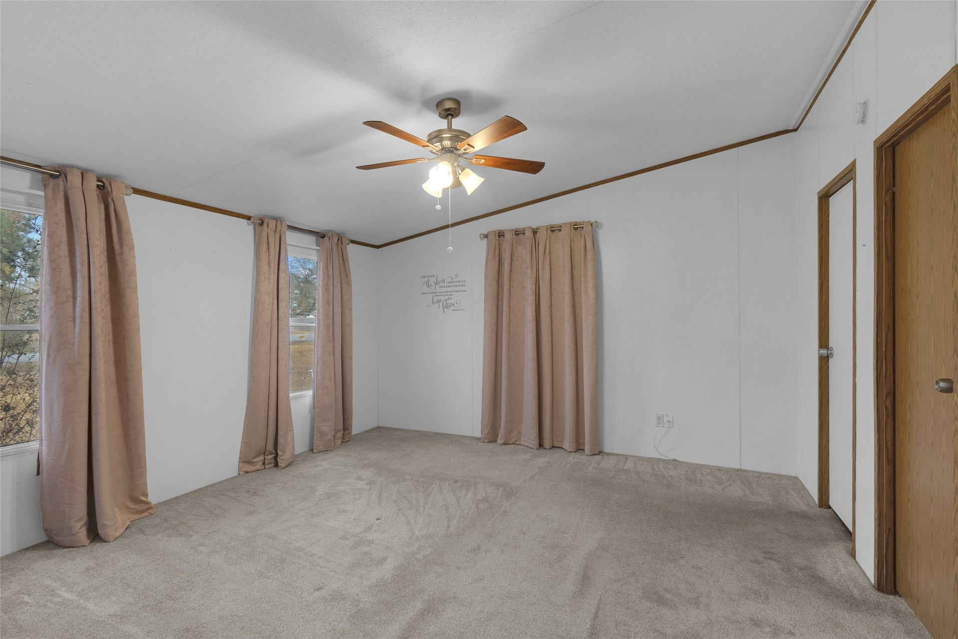 This primary bedroom offers a generous carpeted space with white walls and a ceiling fan. Sunlight streams through the large window with patterned curtains, enhancing the airy and welcoming atmosphere.