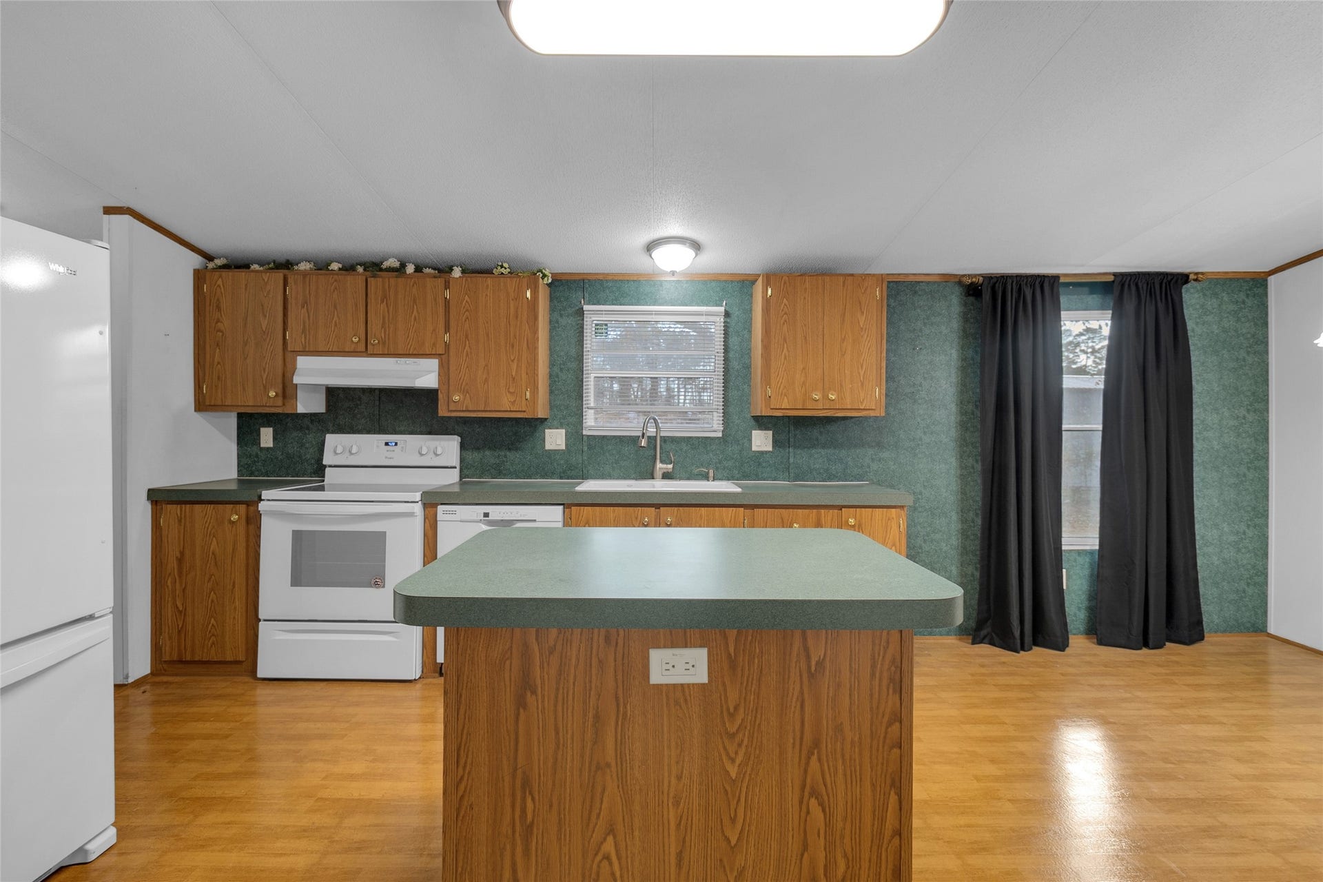 This photo shows a cozy kitchen with wood cabinetry. It features an island, white appliances, and a window over the sink. The room is well-lit and has wood flooring, creating a warm and inviting atmosphere.